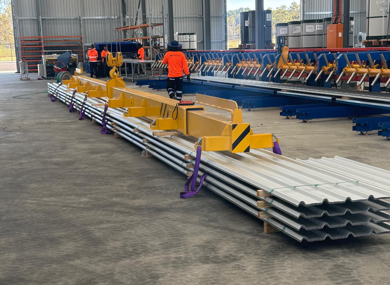 Inside a warehouse or factory, workers are assembling or maintaining large metal roofing sheets. There are several metal sheets, industrial equipment, and workers in high-visibility clothing working on or near machinery.
