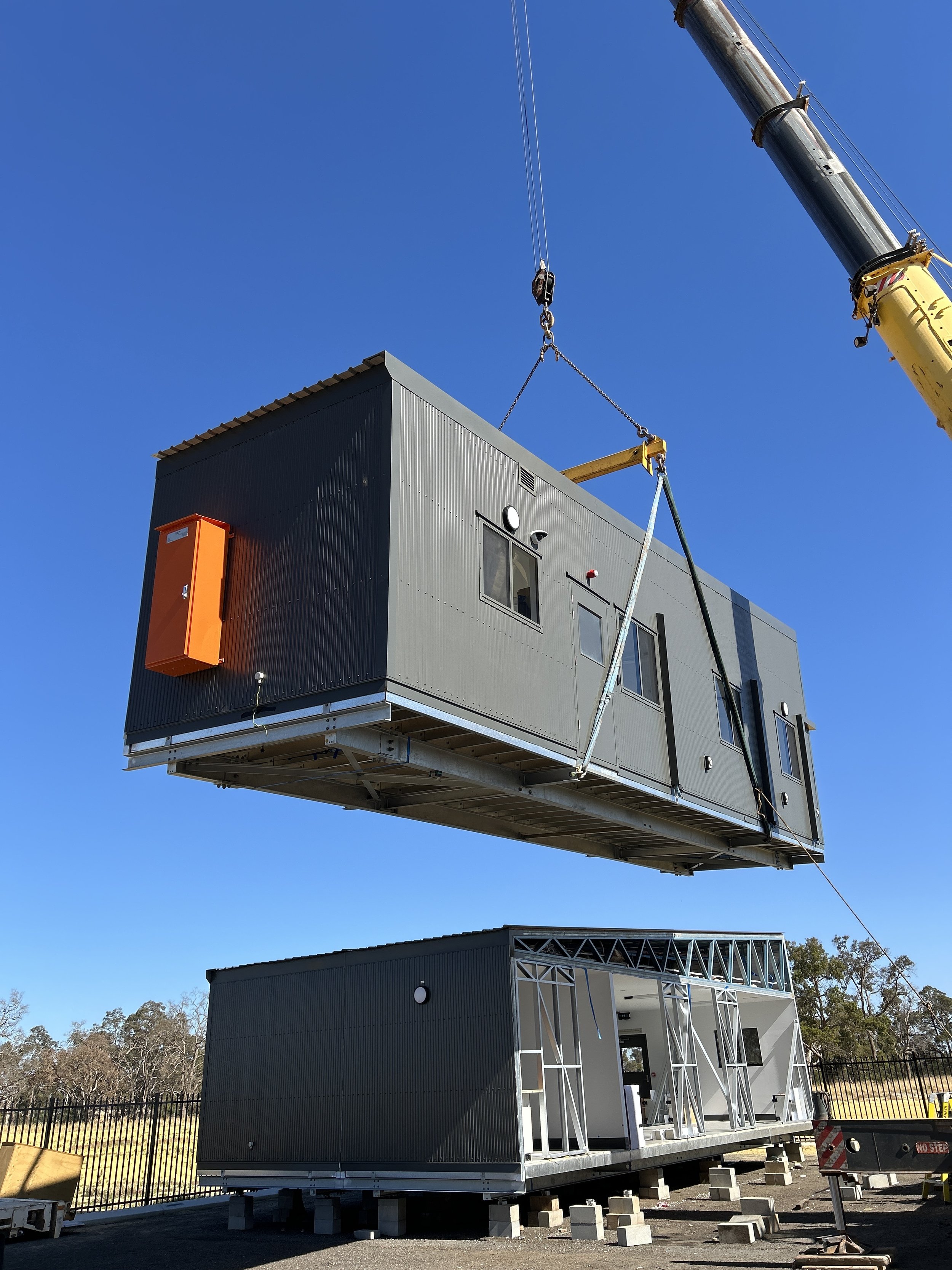 Construction site with modular building being lifted by crane, with a blue sky and trees in the background.