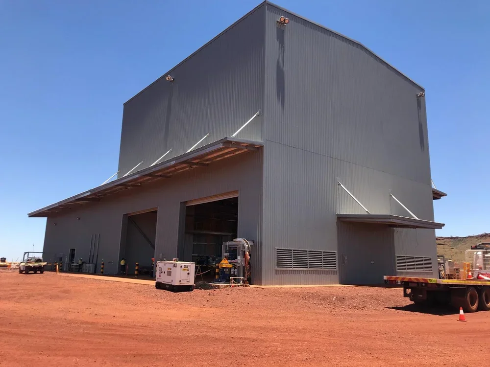 Large industrial warehouse building with metal siding, open garage doors, situated on a dirt lot, under a clear blue sky.