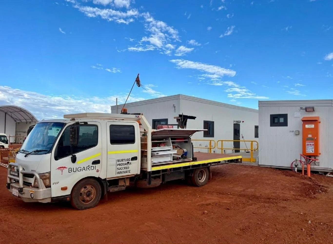A white utility truck with equipment in the back, parked on reddish dirt ground near small white buildings with black windows, under a partly cloudy blue sky.