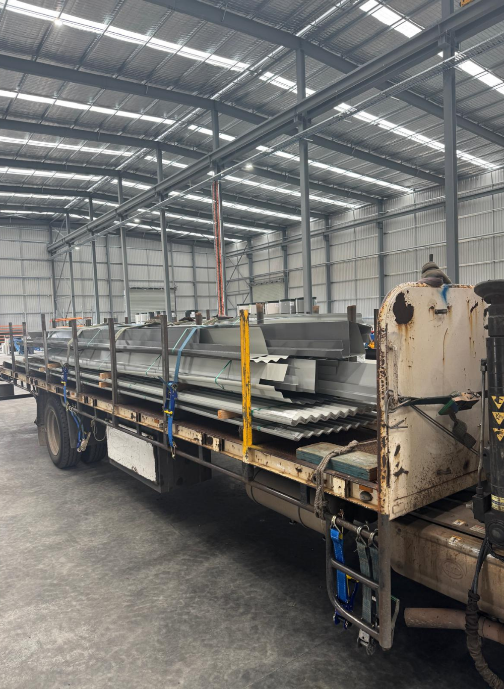 A flatbed truck inside a large industrial warehouse, carrying metal panels and sheets secured with straps, with a high ceiling and steel beams overhead.