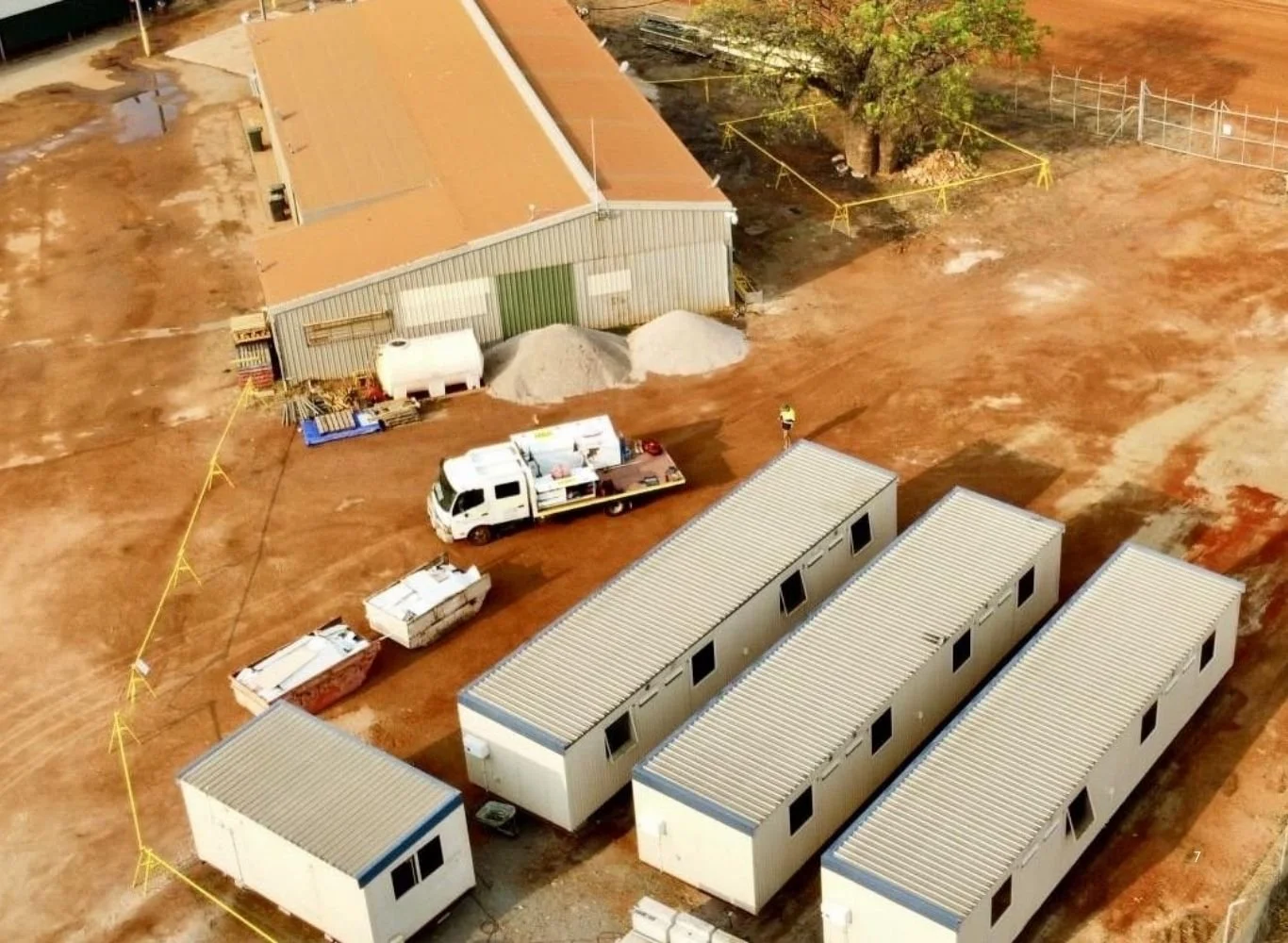 Aerial view of a construction site with portable buildings, a large metal warehouse, construction equipment, and dirt ground.