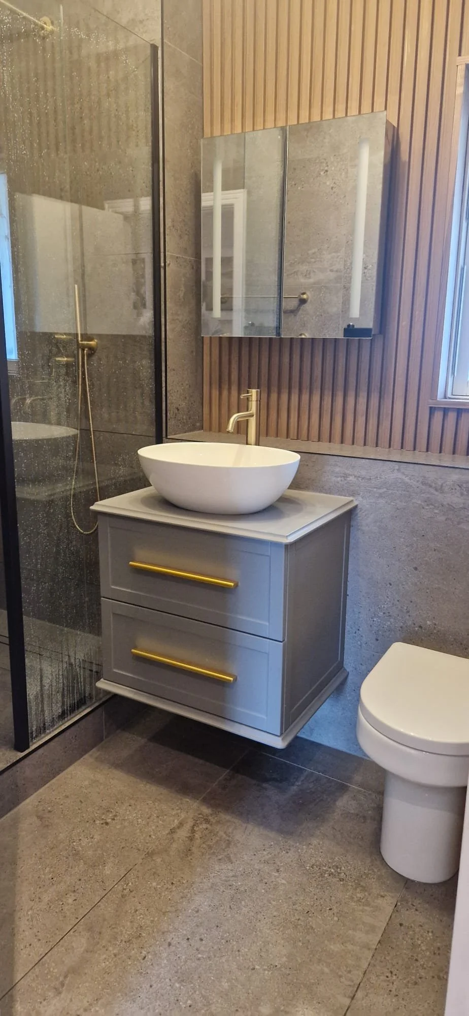 Modern bathroom with a floating vanity, vessel sink, gold faucet, and a large mirror. There is a glass shower enclosure with a gold showerhead on the left side and a window on the right wall. The walls are partly covered in wood paneling and stone ti