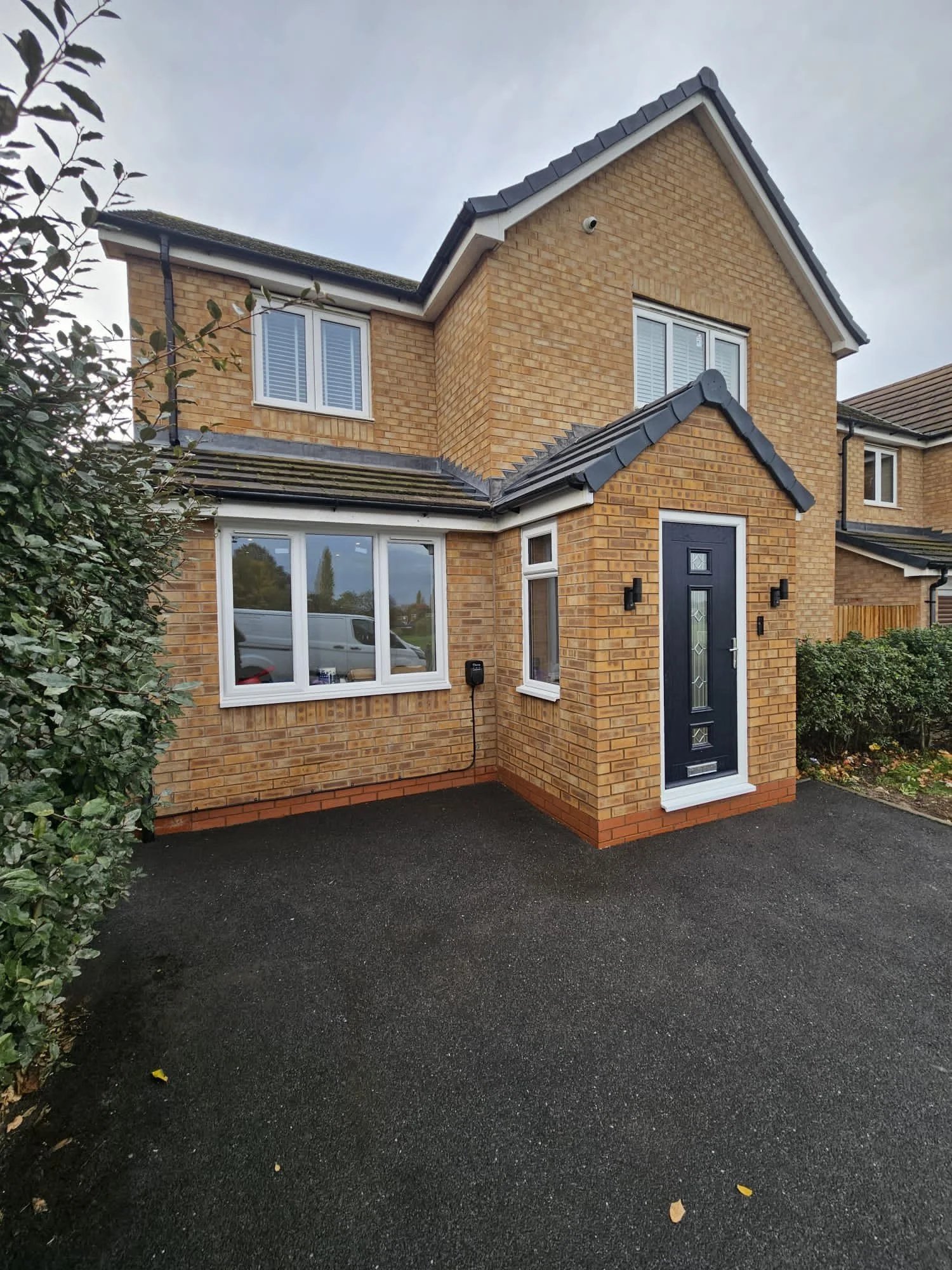 front view of a two-story brick house with a black front door and white window frames, a large window on the ground floor, two on the upper floor, and a paved driveway.