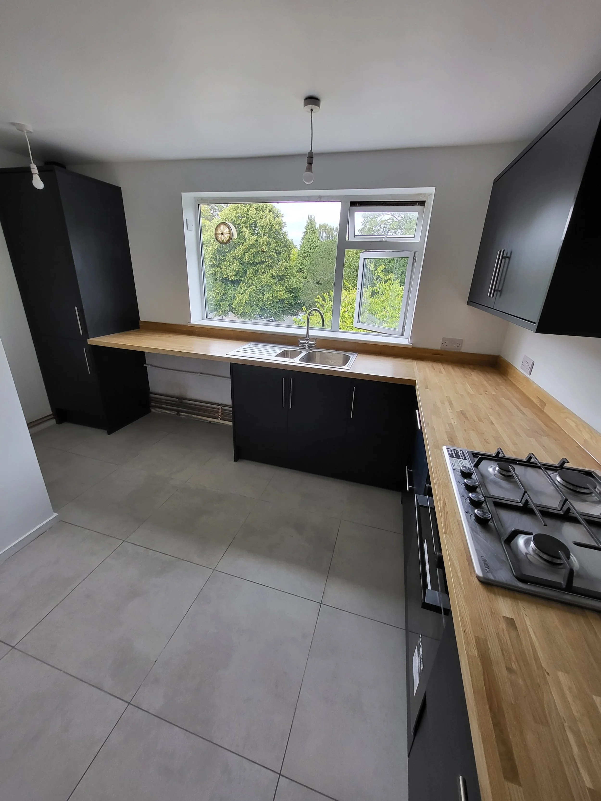 Modern kitchen with black cabinets, wooden countertops, a double sink under a large window showing greenery outside, and a gas stove on the right. The room has light-colored tile flooring.
