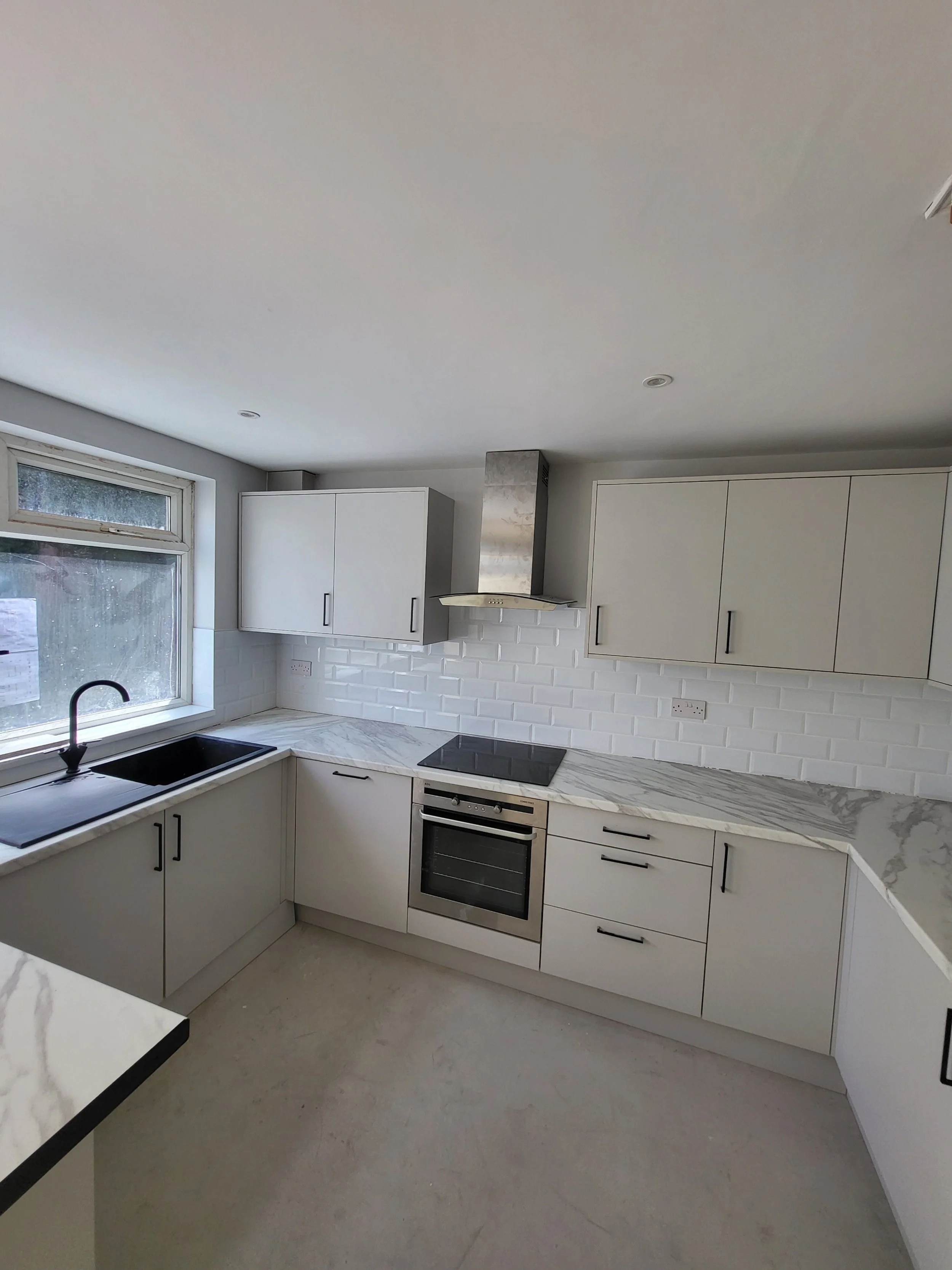 Modern kitchen with white cabinets, marble countertops, black handles, stainless steel oven, black induction cooktop, stainless steel range hood, black sink, and a window