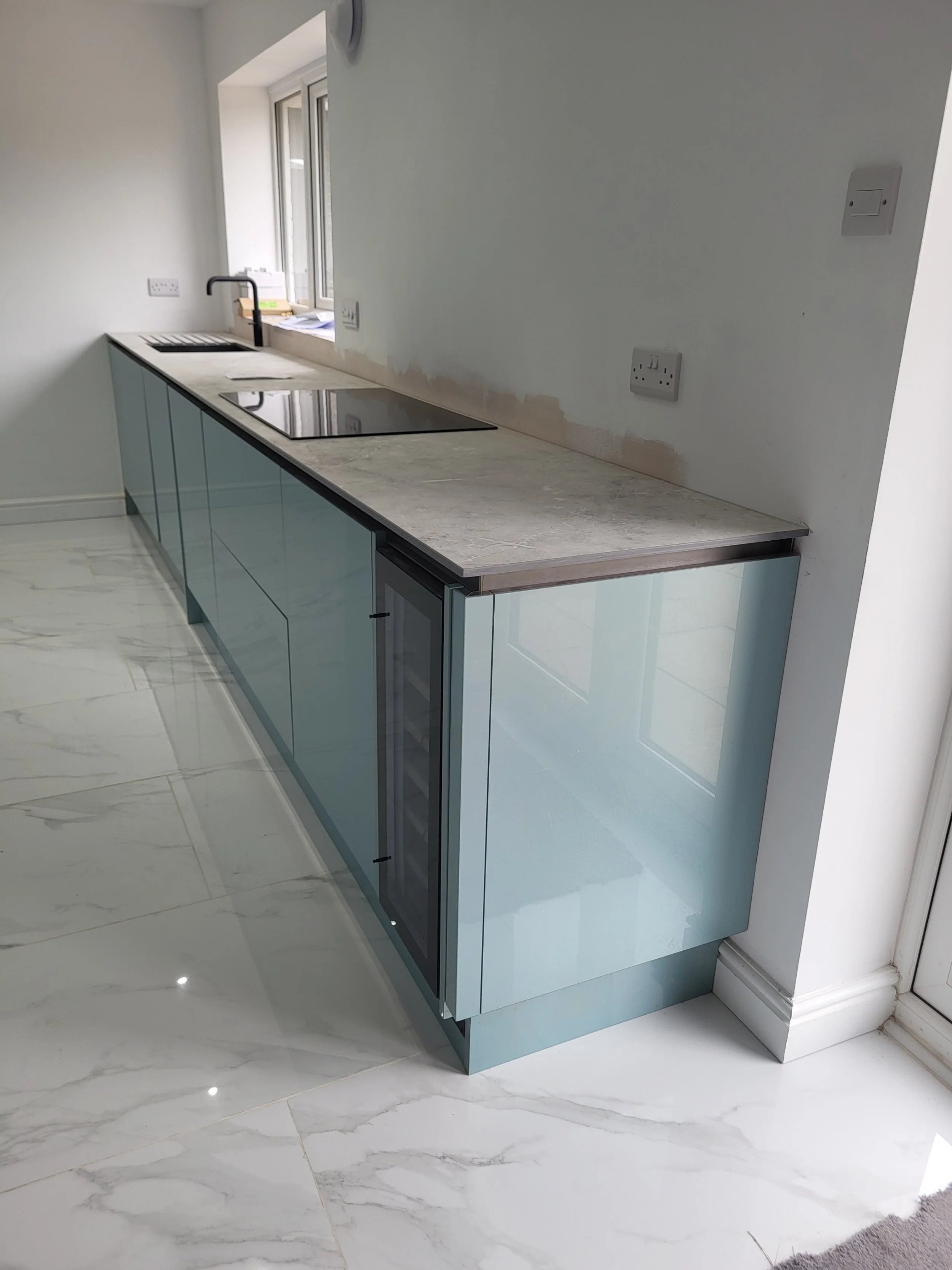 Modern kitchen with light blue cabinets, marble floor, and a countertop with a sink, electric cooktop, and window above.