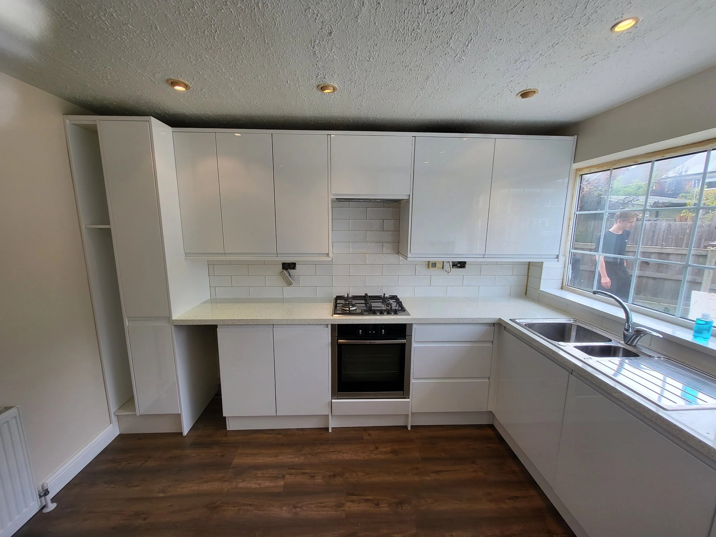 Kitchen with white cabinets, white countertop, gas stove, and a large window overlooking the backyard with a person outside.