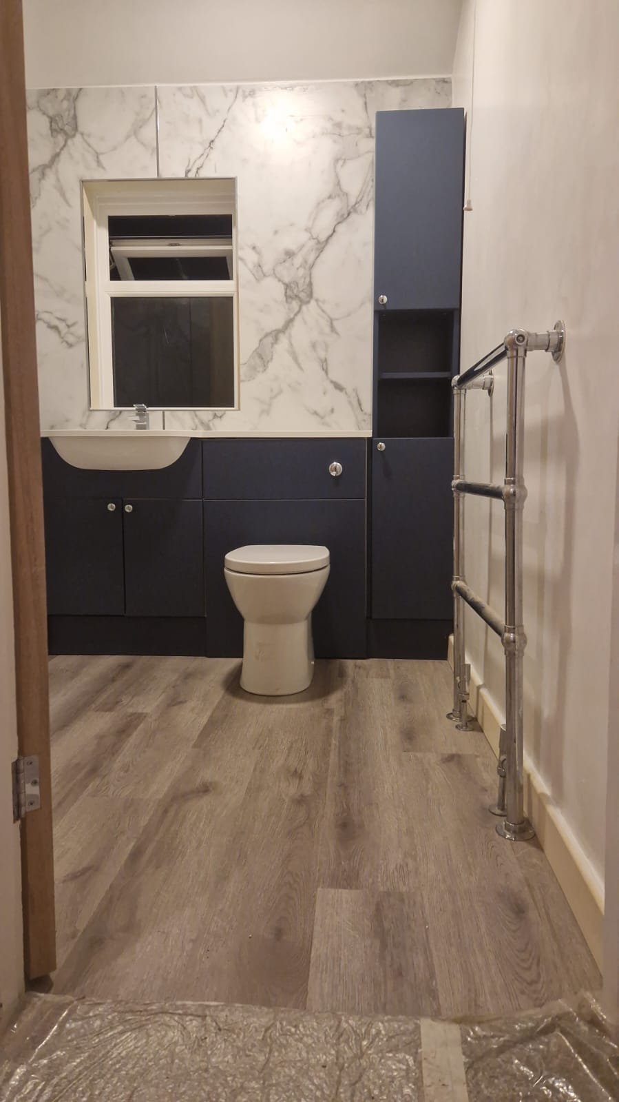 Bathroom with marble wall, small mirror, dark blue cabinet, white toilet, metal towel rack, wooden flooring, and partial door frame.