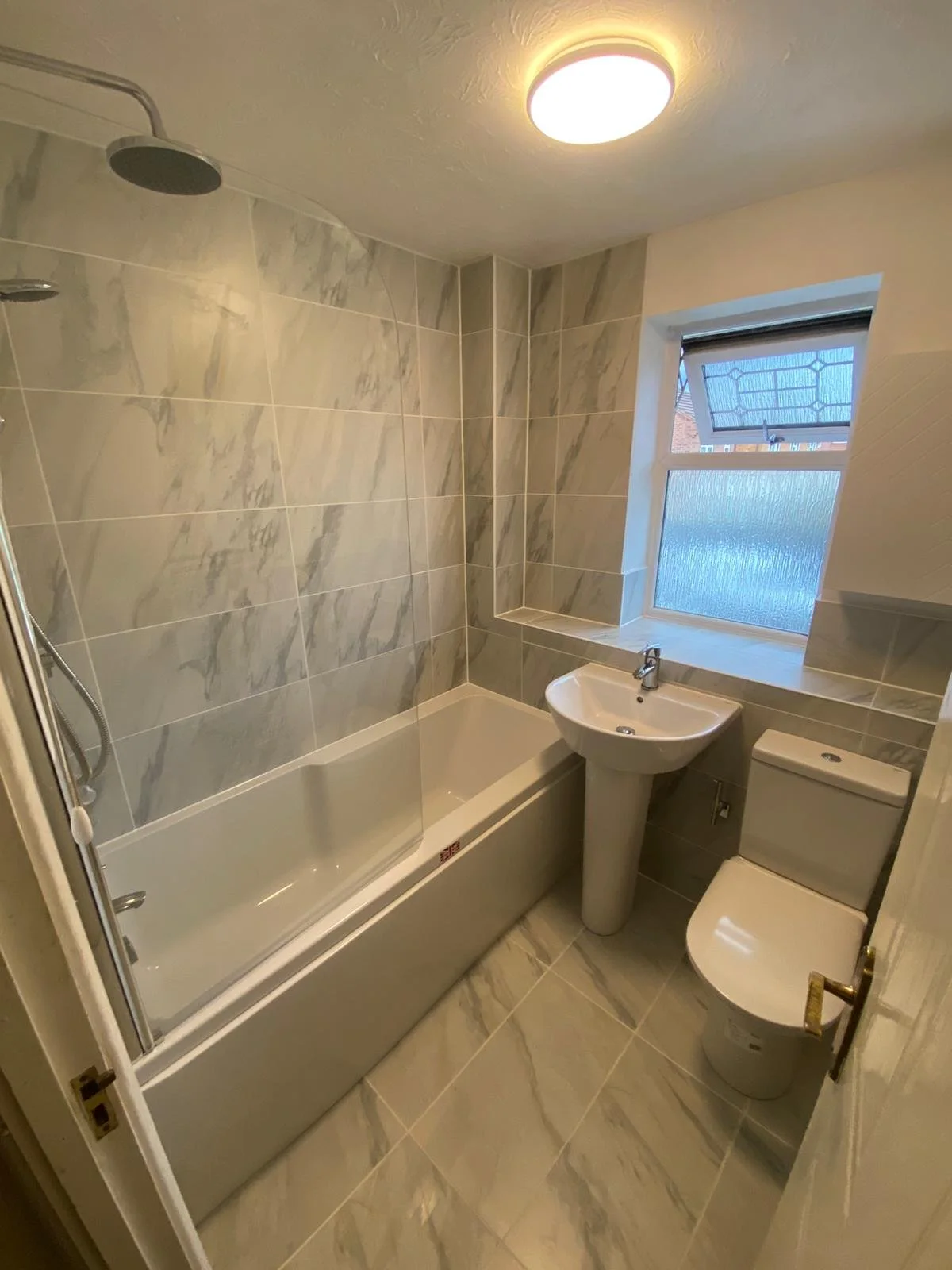 Small bathroom with a bathtub, a pedestal sink, a toilet, a frosted window, and marble-patterned tiles on the walls and floor.