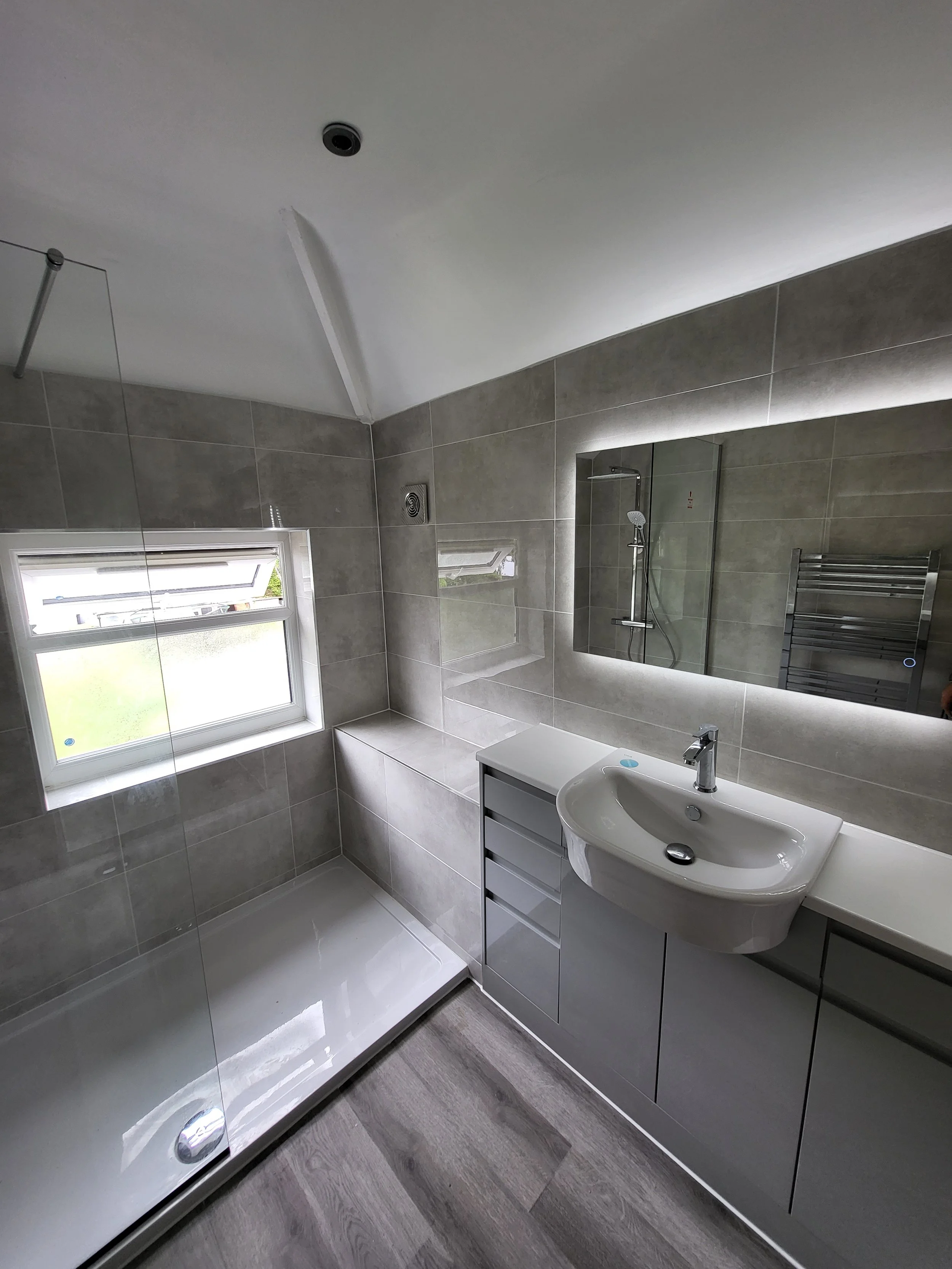 Modern bathroom with gray tiled walls, a walk-in shower with a glass partition, a white vanity with a curved sink, a large mirror with backlighting, a small window, and wood-look flooring.