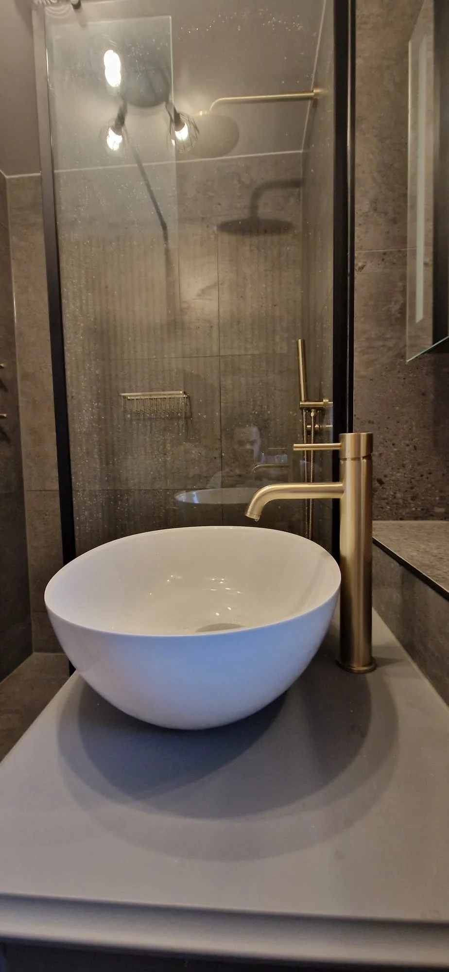 Close-up of a modern bathroom vanity with a white vessel sink, brushed gold faucet, and a mirror reflecting a person taking the photo.