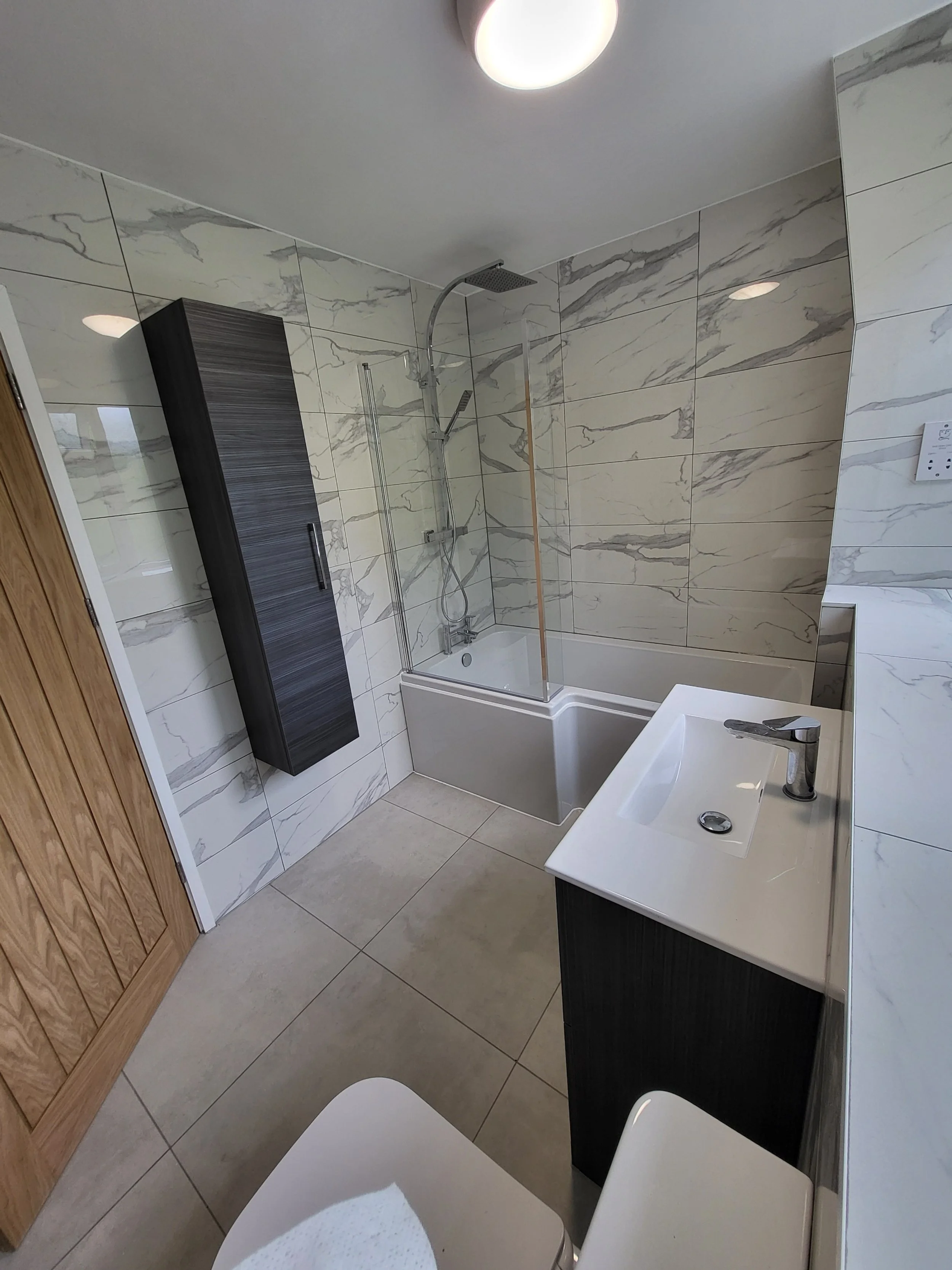 A modern bathroom with marble tiles on the walls, a bathtub with a glass shower screen, a dark wood cabinet mounted on the wall, a white sink with a dark cabinet underneath, a toilet, and a wooden door. There is a ceiling light fixture.