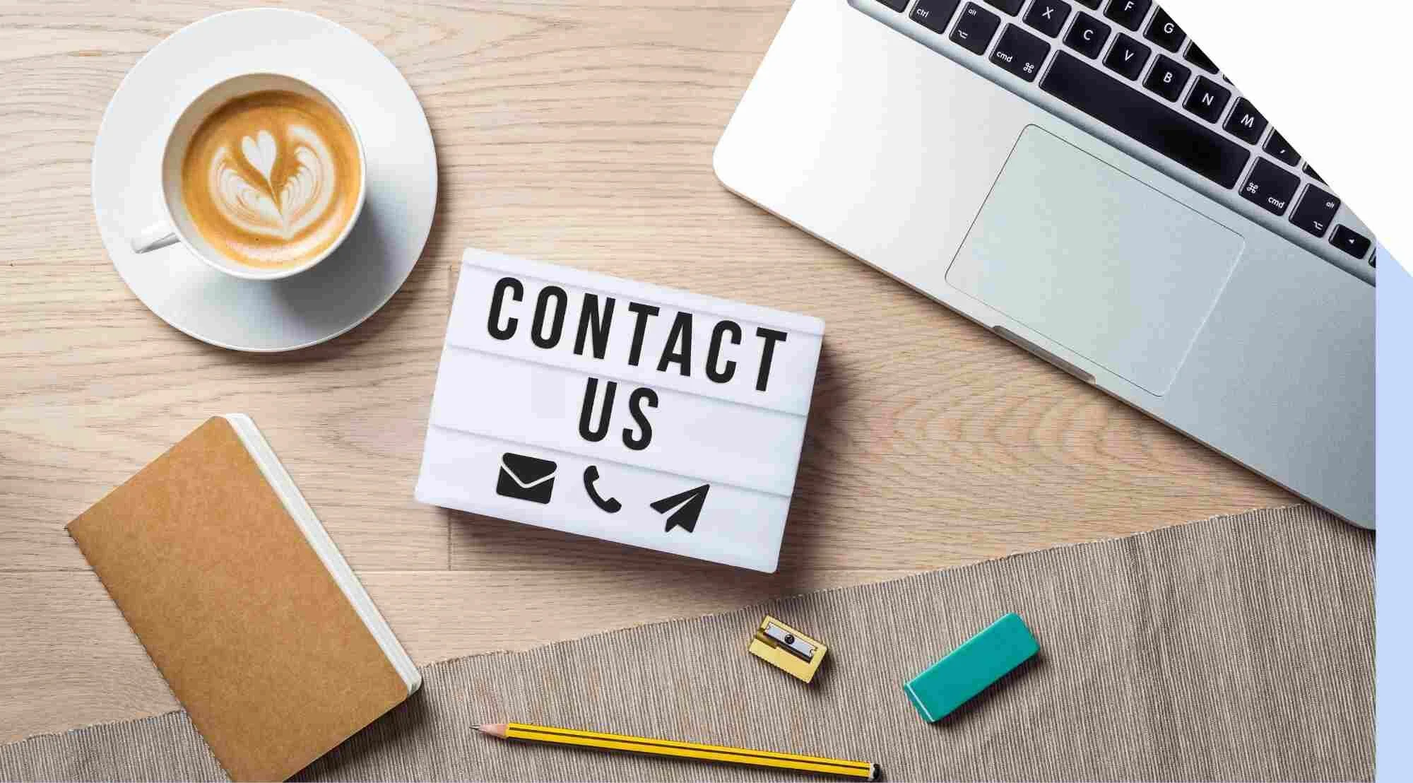 Top-down view of a workspace with a cup of latte, a small lightbox sign saying 'CONTACT US' with icons for email, phone, and messaging, a laptop, a notebook, a pencil, a small sharpener, and an eraser on a desk.