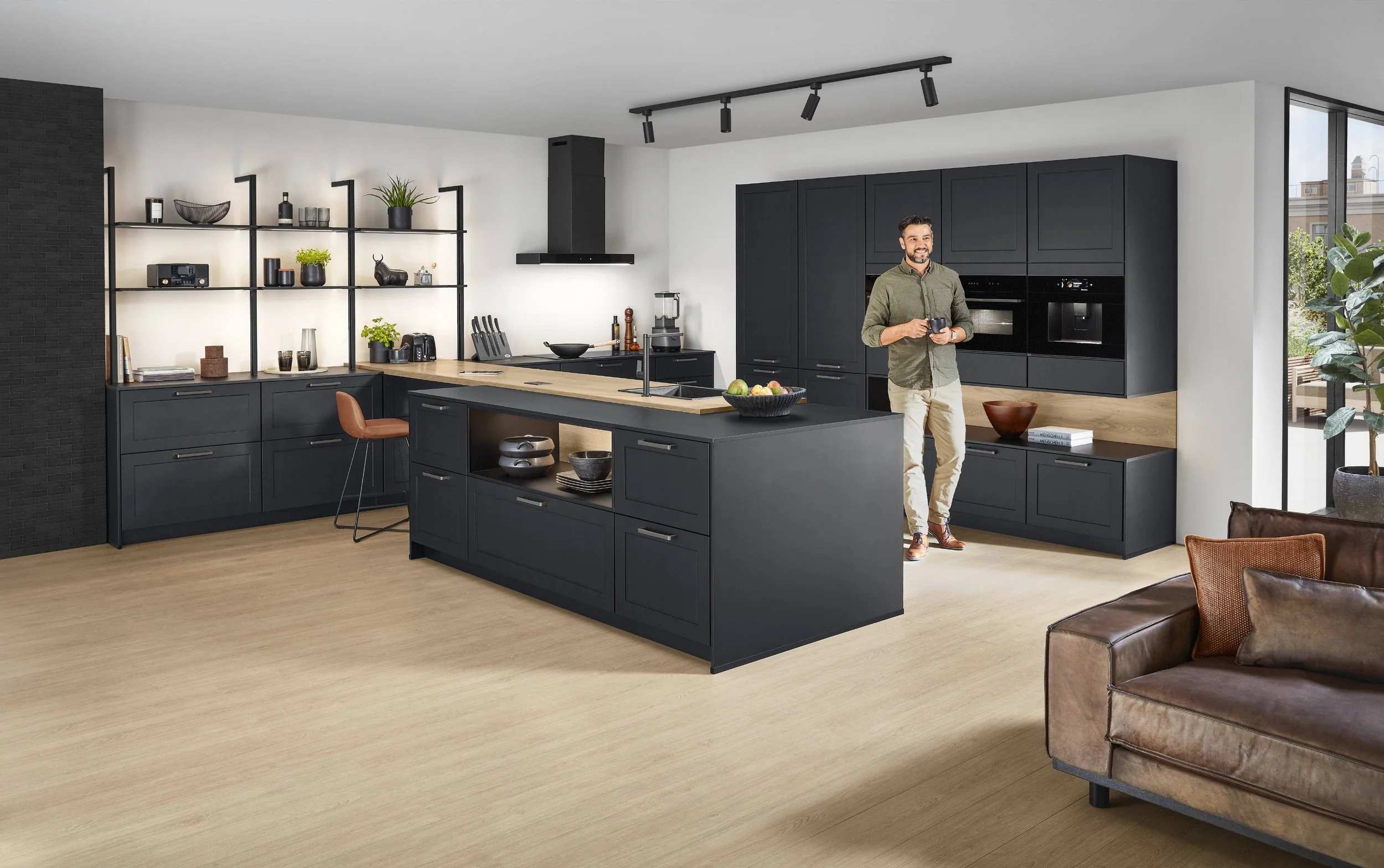Moderne offene Küche mit schwarzen Schränken, Kücheninsel und Holzarbeitsflächen, eine Person hält eine Tasse, großer Wohnbereich mit Sofa und viel Tageslicht.