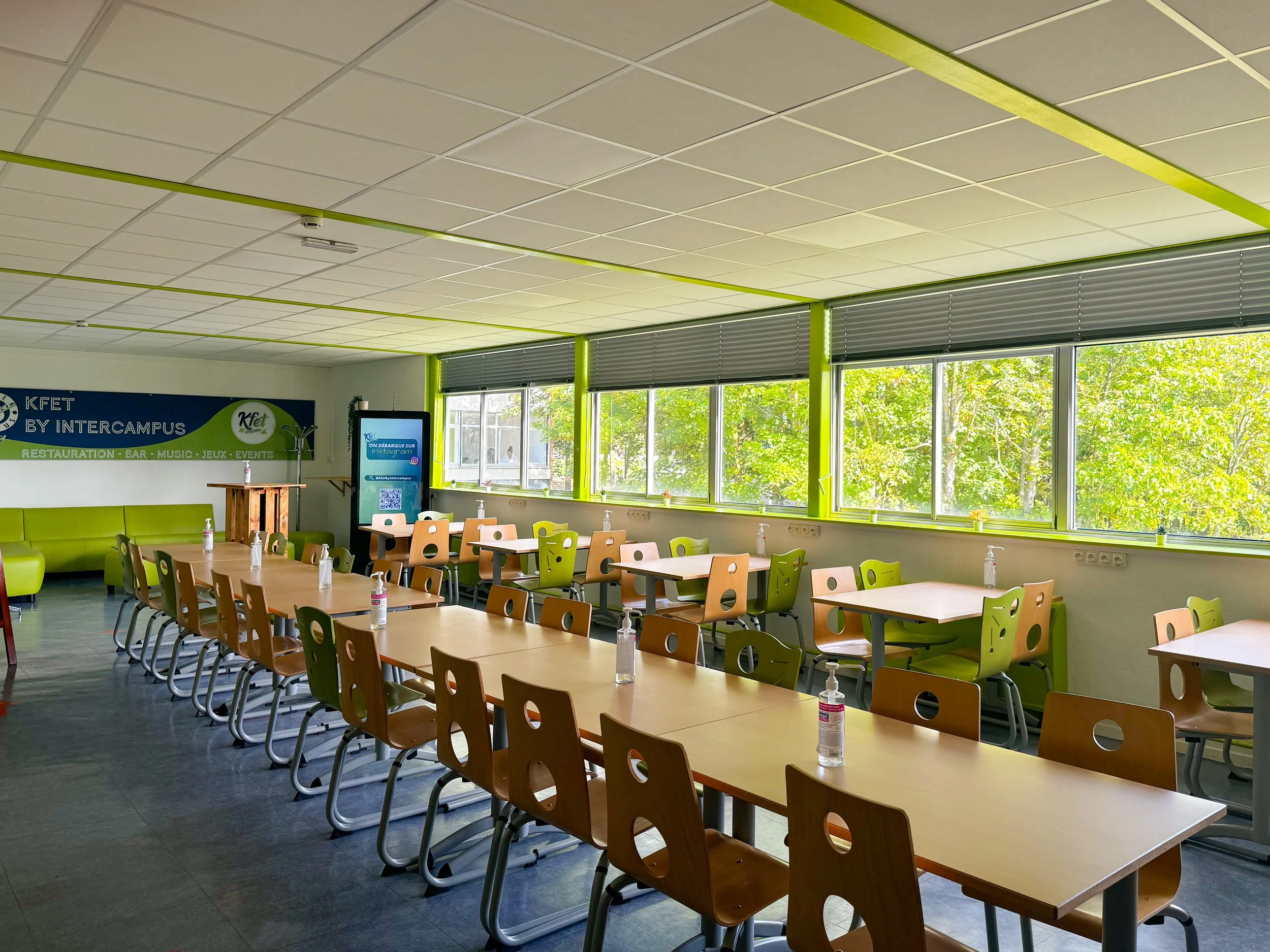 Salle de réunion avec des tables en bois, des chaises en plastique et des bouteilles de gel désinfectant. Grande fenêtre laissant entrer de la lumière naturelle, vue sur des arbres verdoyants. Mur avec affiches et un écran numérique.
