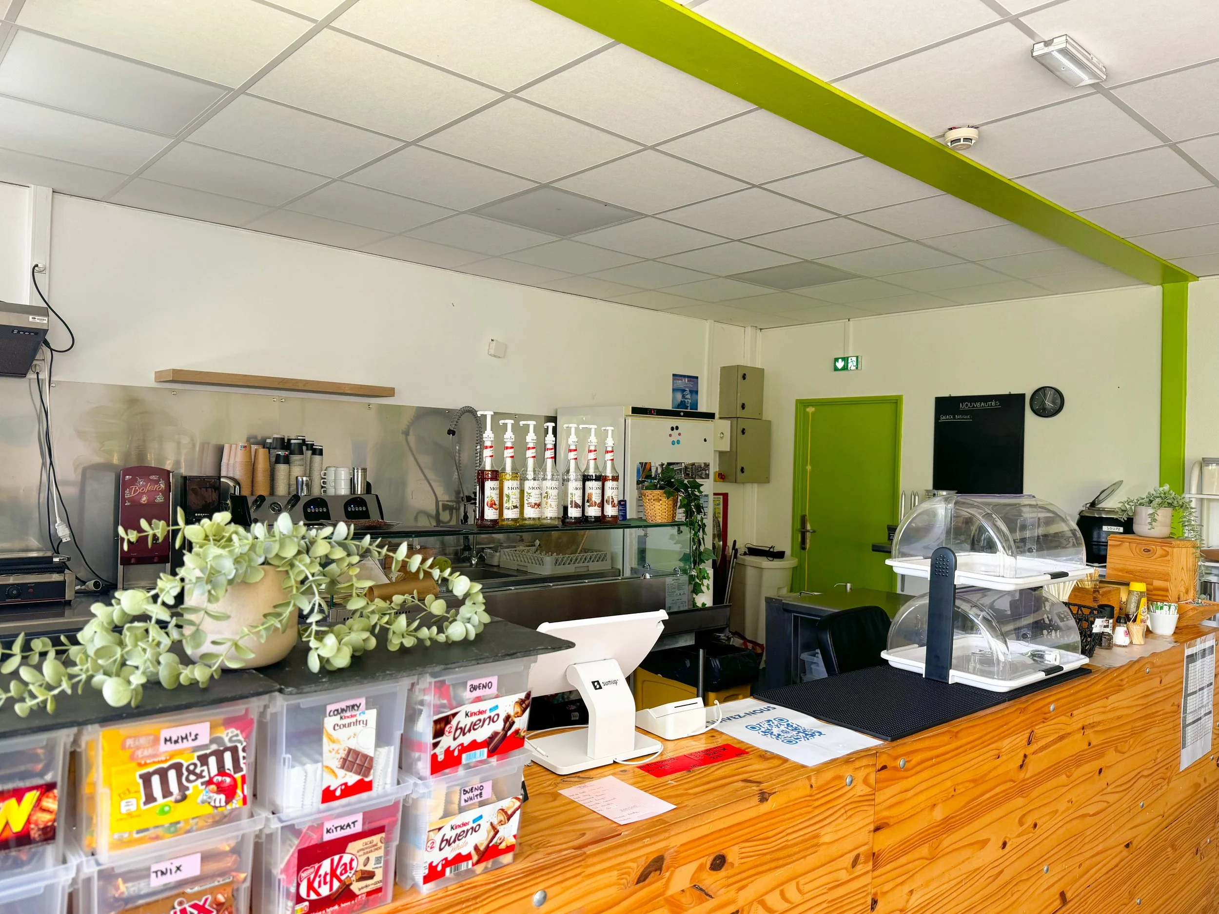 Une image d’un comptoir dans un café ou un snack, avec des boissons, des confiseries et des accessoires sur le comptoir en bois, et un mur blanc avec un panneau noir indiquant des nouveautés et une horloge.