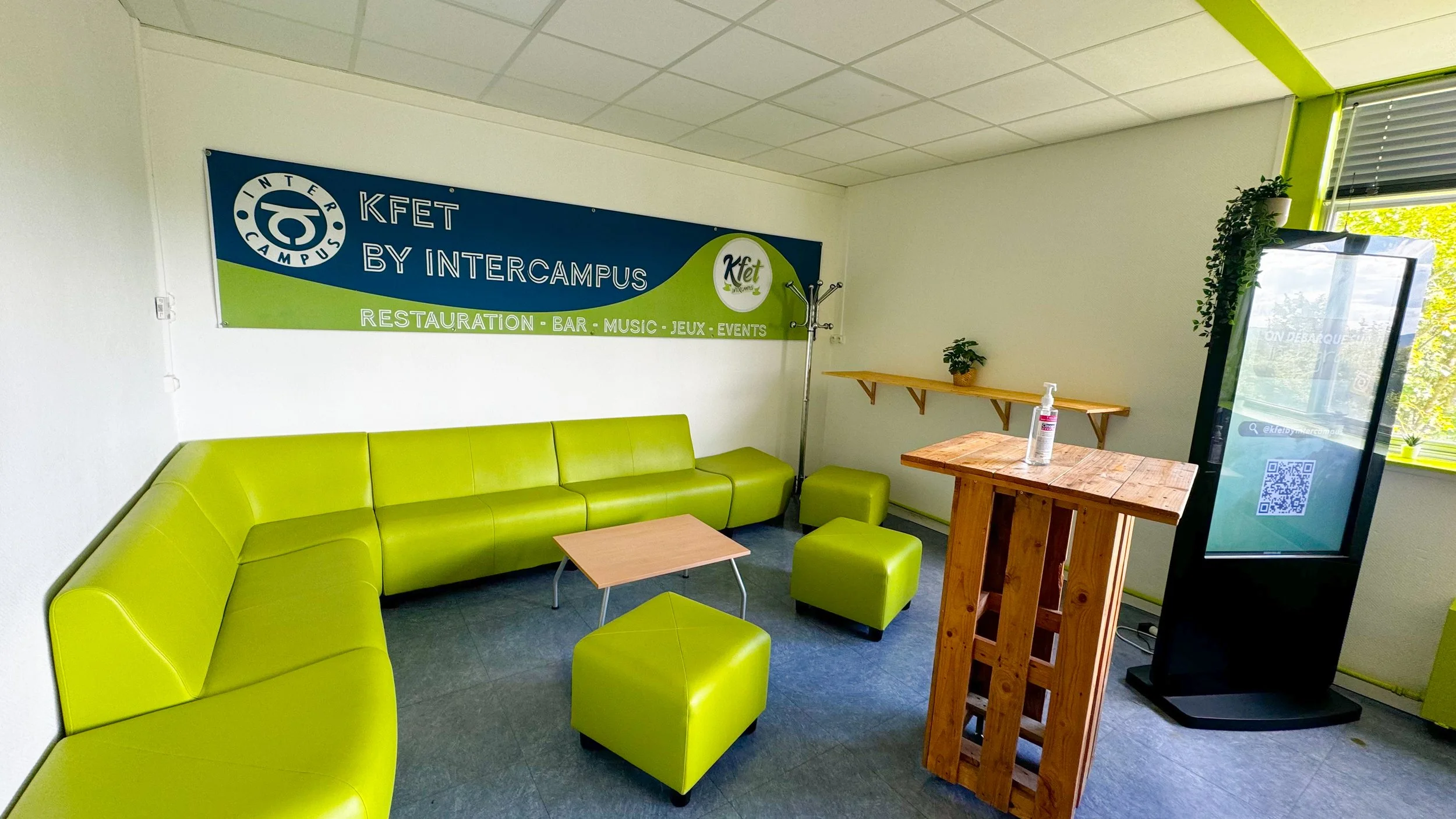 Intérieur d'un espace lounge avec canapé vert en U, petites tables et plantes vertes, panneau publicitaire de l'Intercampus KFET, fenêtre laissant entrer la lumière naturelle.
