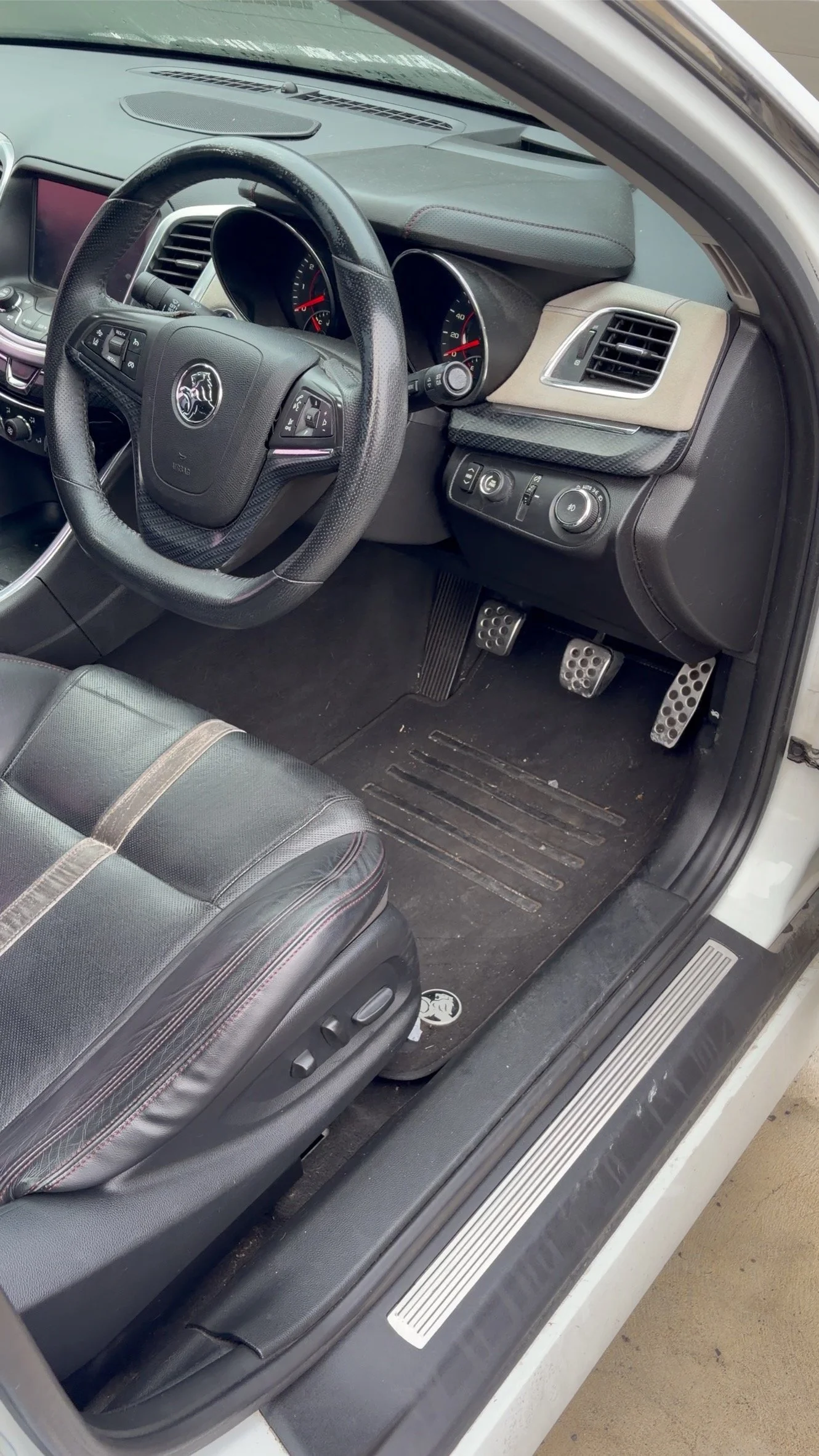 Inside a car, showing the driver's seat, steering wheel with a Cadillac logo, dashboard with speedometer, air vent, and control switches, along with pedals and a floor mat.