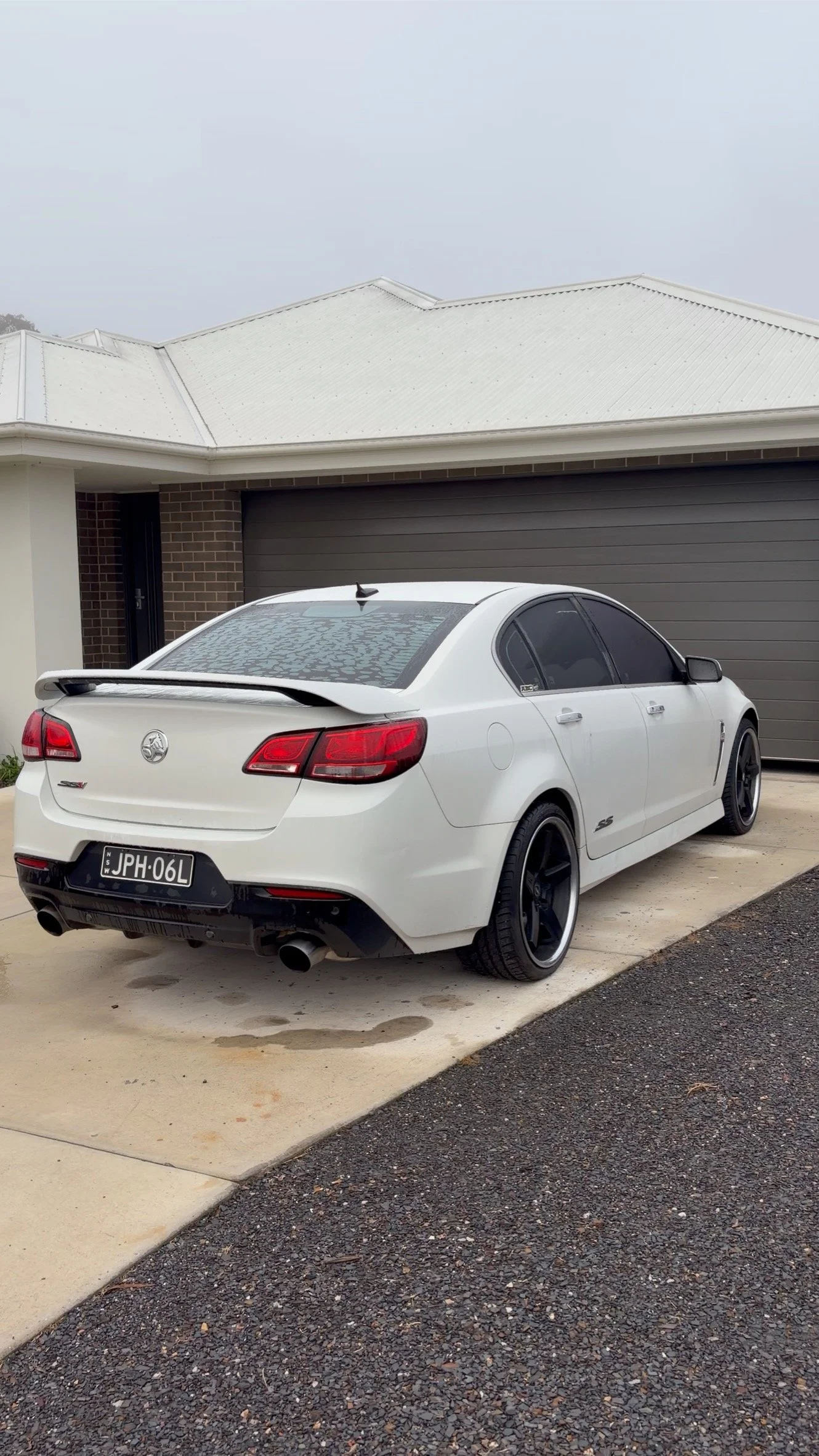 White sedan car with a rear spoiler and tinted windows parked in front of a modern house with a grey garage door and white roof, on a driveway with concrete and gravel surfaces.