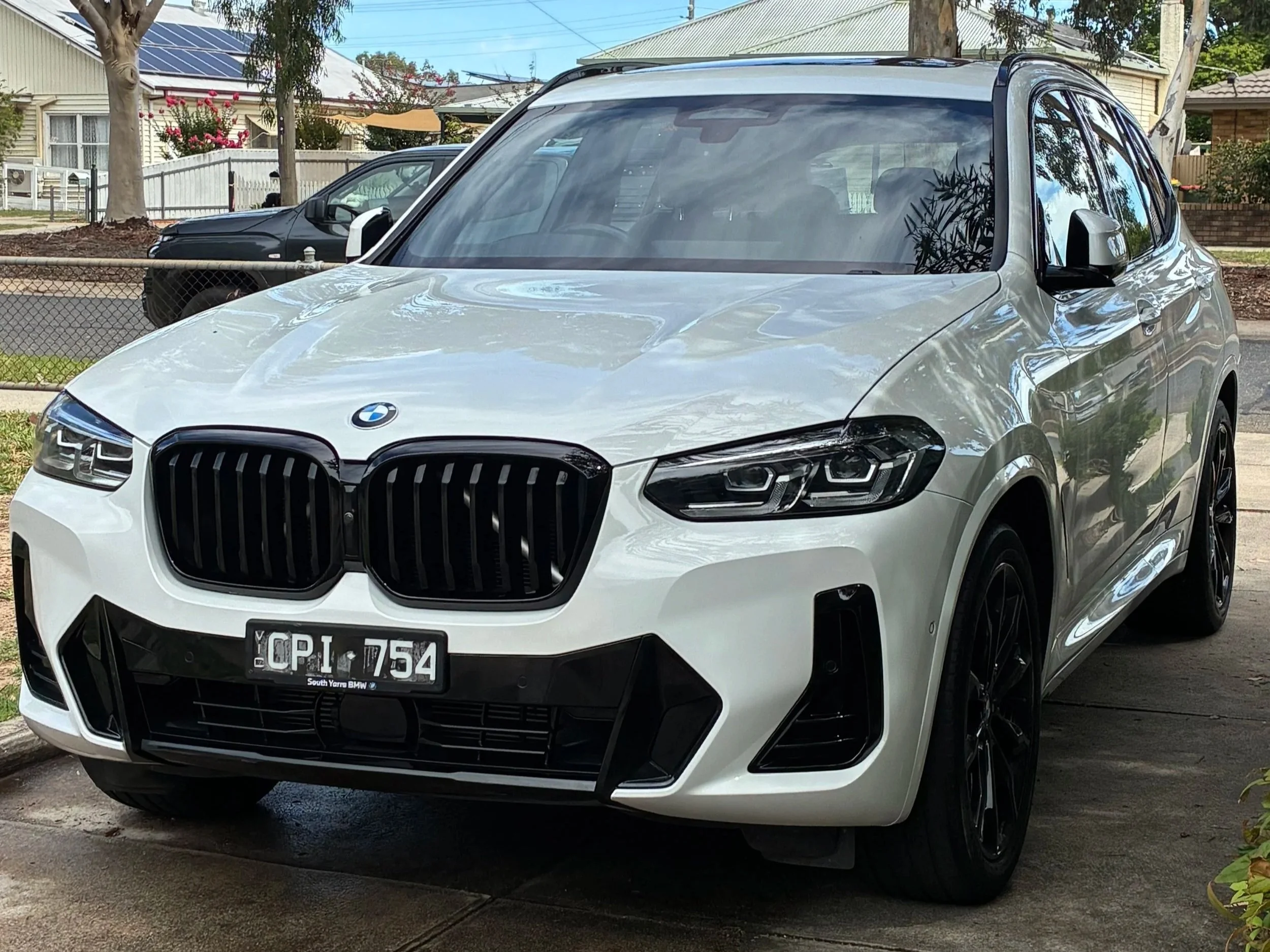 A white BMW SUV parked on a driveway in a suburban neighborhood, with another black vehicle in the background and trees and houses nearby.