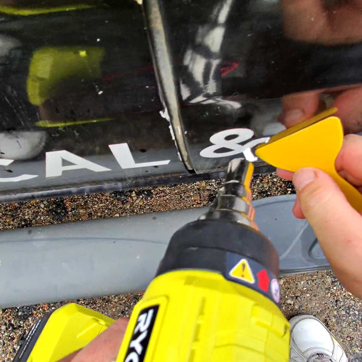 Person using a power drill to remove a white letter 'A' from a black surface of a vehicle or object.