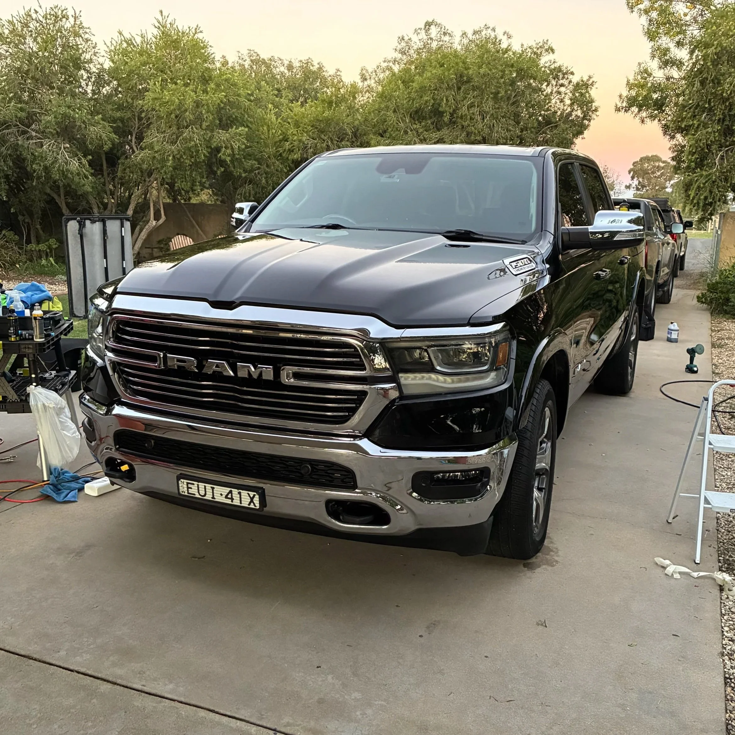 A black RAM pickup truck parked on a driveway with equipment and tools around it, trees in the background, and a sunset sky.