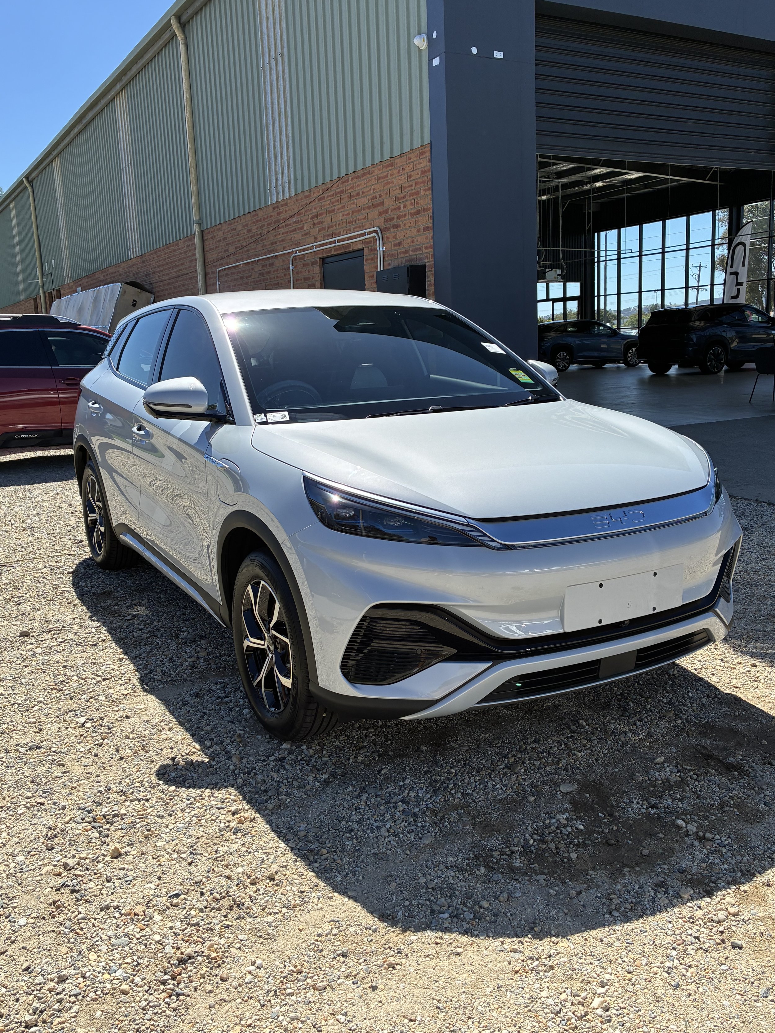 A white electric SUV parked outside a car dealership, with a brick and metal building and other vehicles in the background.