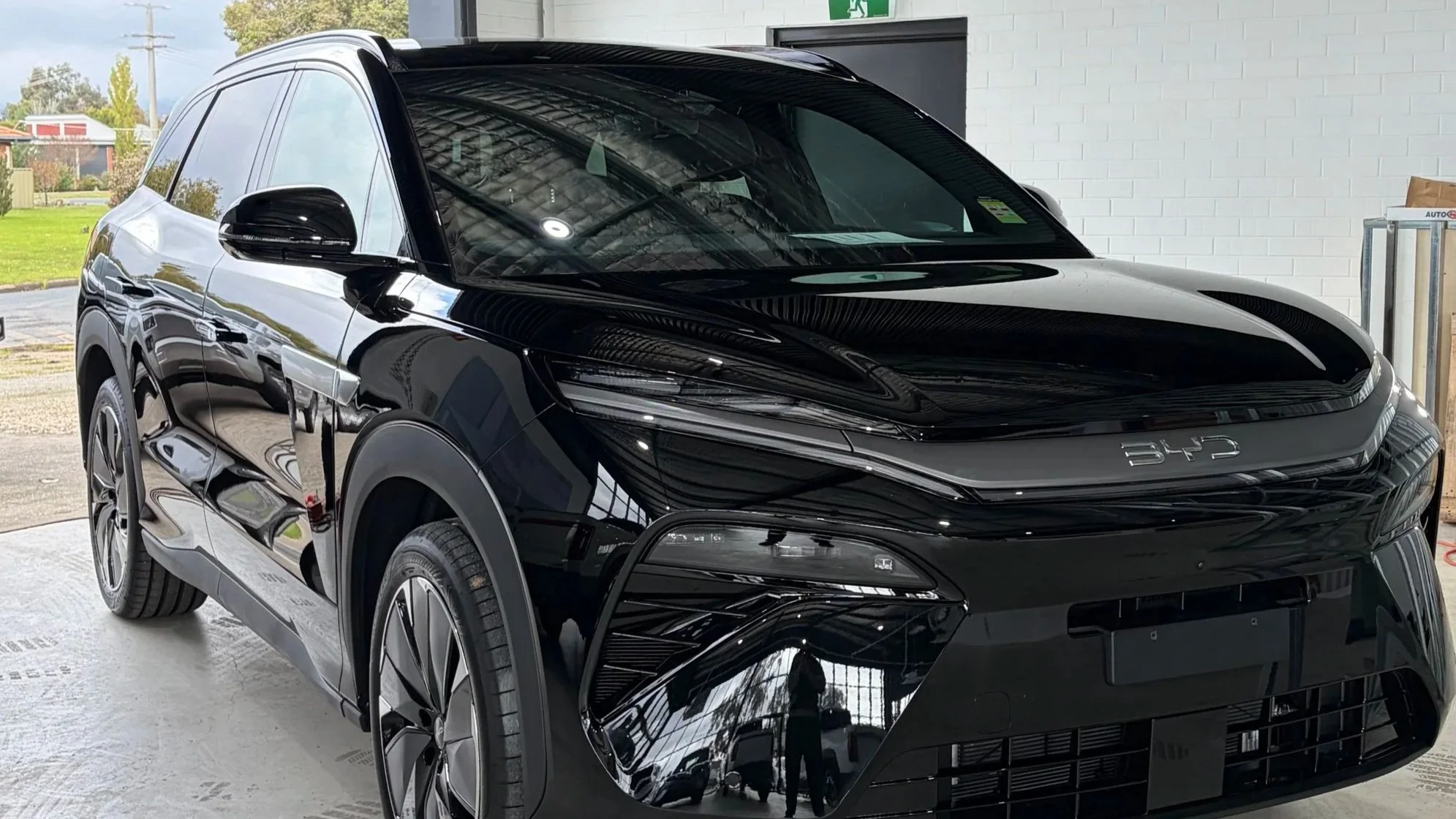 Black electric SUV parked indoors with a white brick wall and a partially visible green emergency exit sign behind it.