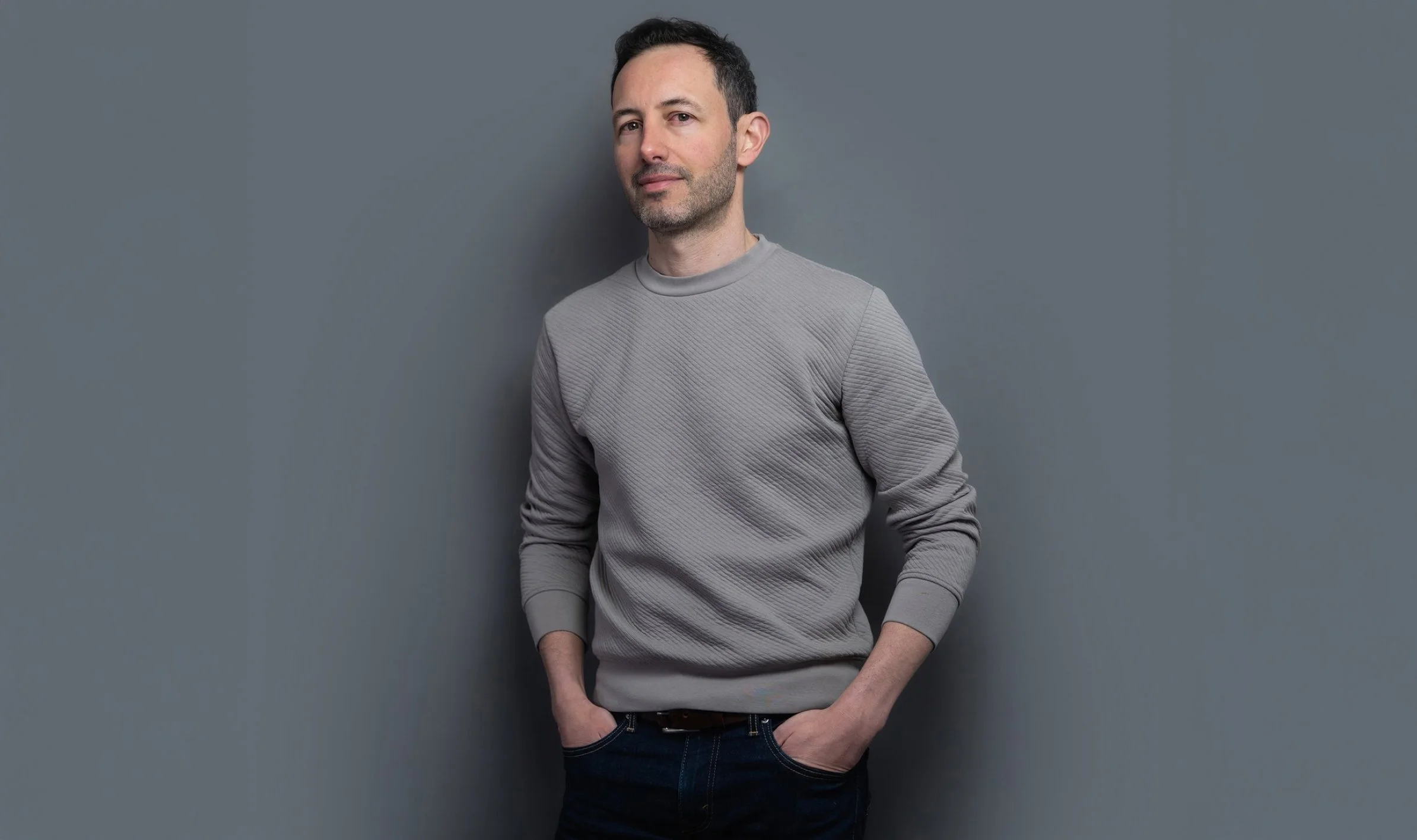 David Cliff is a freelance graphic design based in York. Pictured here standing against a grey wall, wearing a light grey textured sweatshirt and dark jeans, with short dark hair.