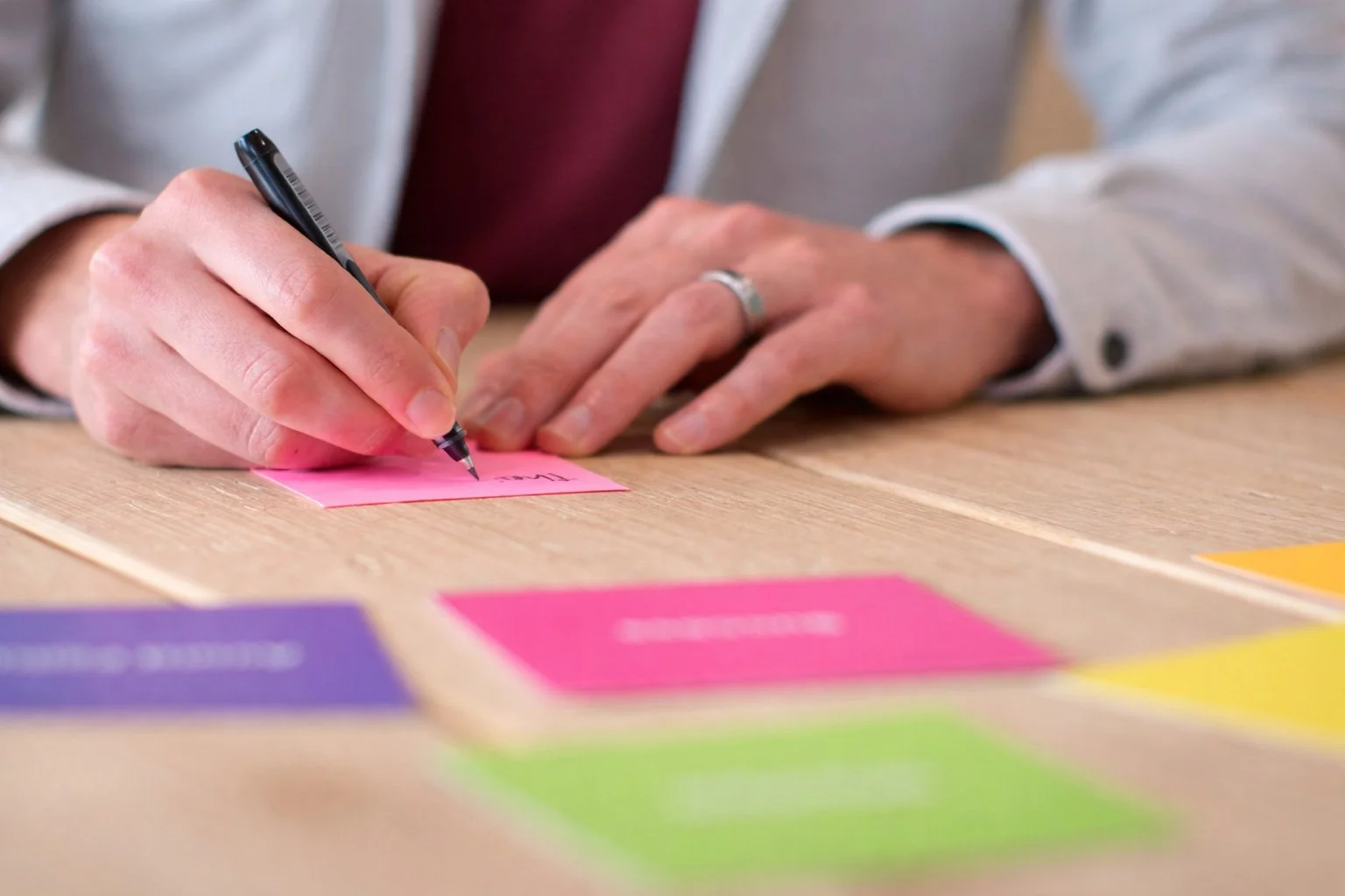 Close up of hands writing on a sticky note with an assortment of brightly coloured prompt cards spread on the desk