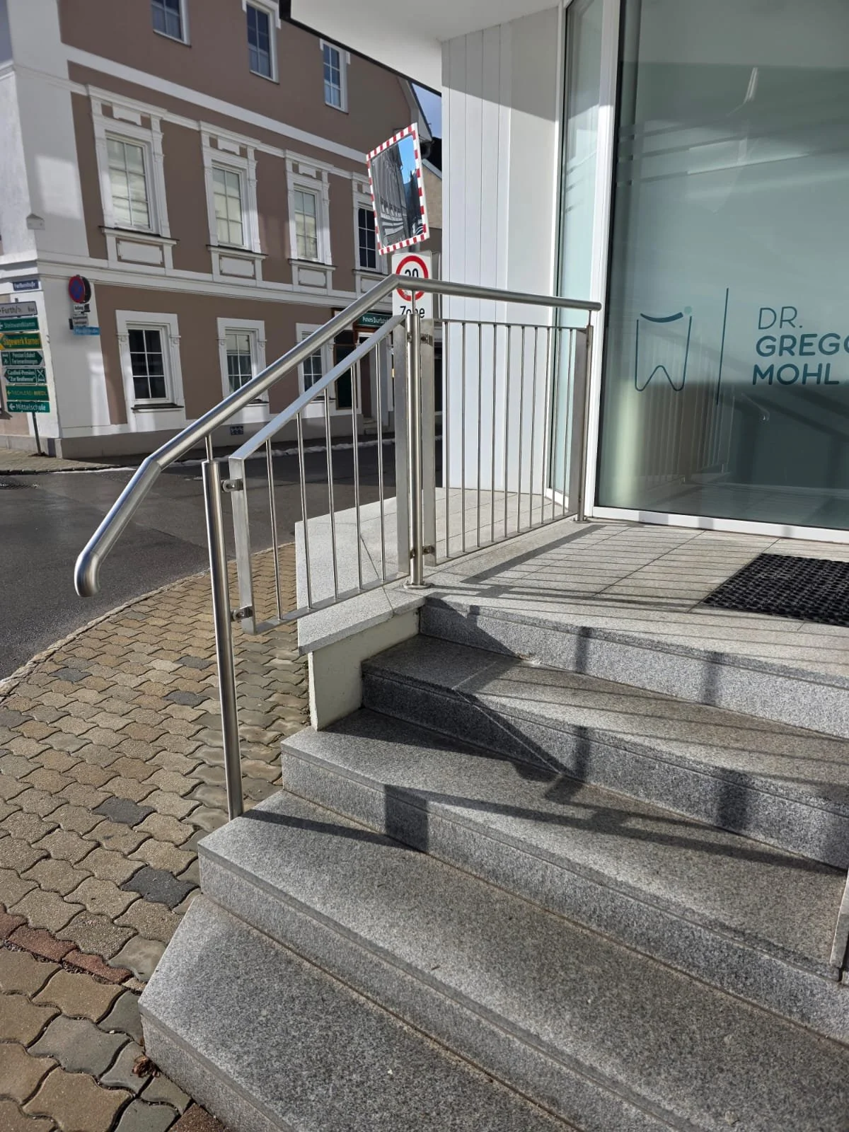 Vorderansicht eines Gebäudes mit einem Zugang, Stufen, Glasfenster und Türen, auf der rechten Seite eine Glasfassade mit einem Zahnsymbol.