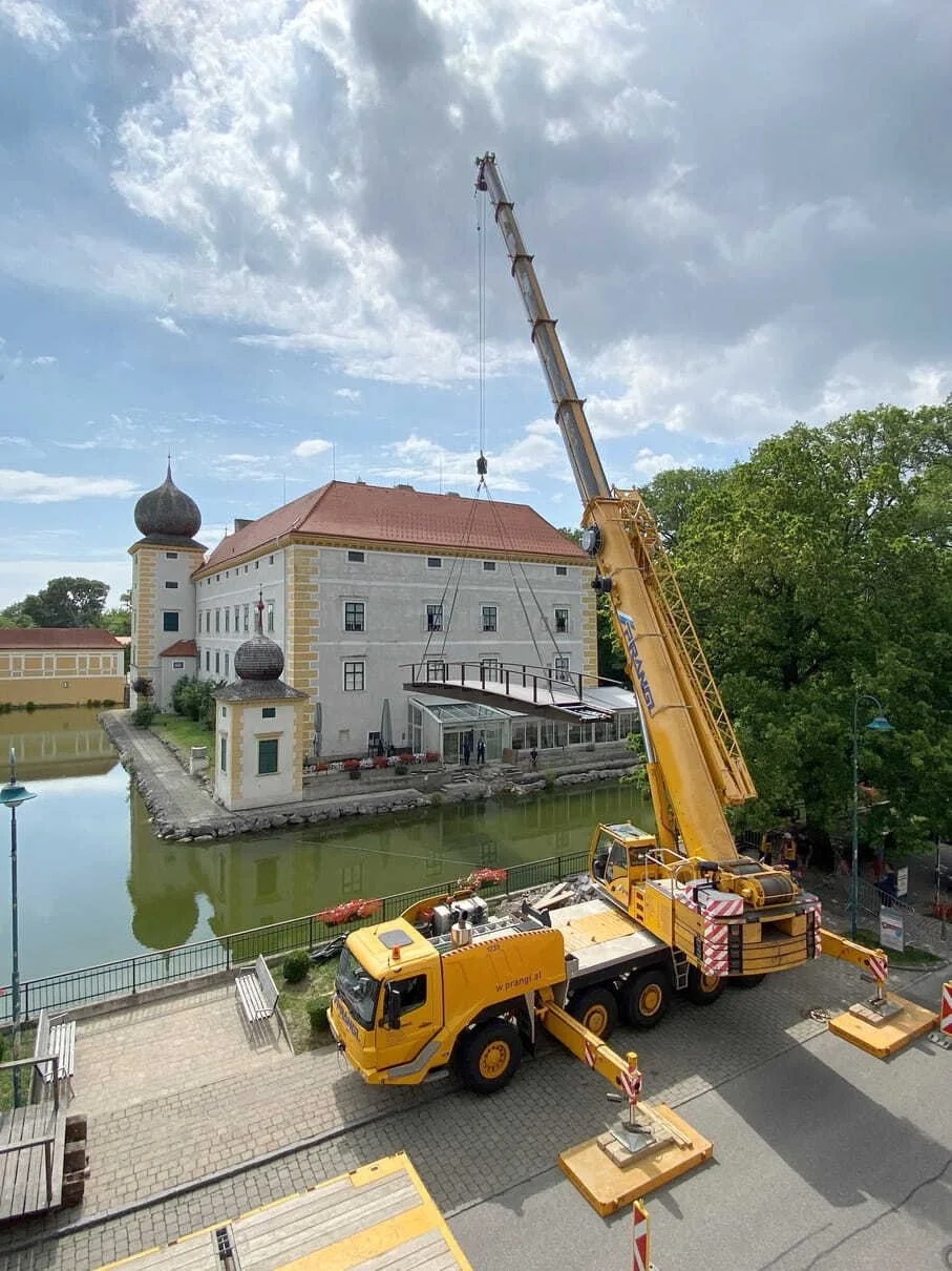 Kran hebt ein Plattformdach vor einem historischen Schloss mit Wassergraben. Das Gebäude hat einen roten Ziegeldach und eine elegante Turmspitze.