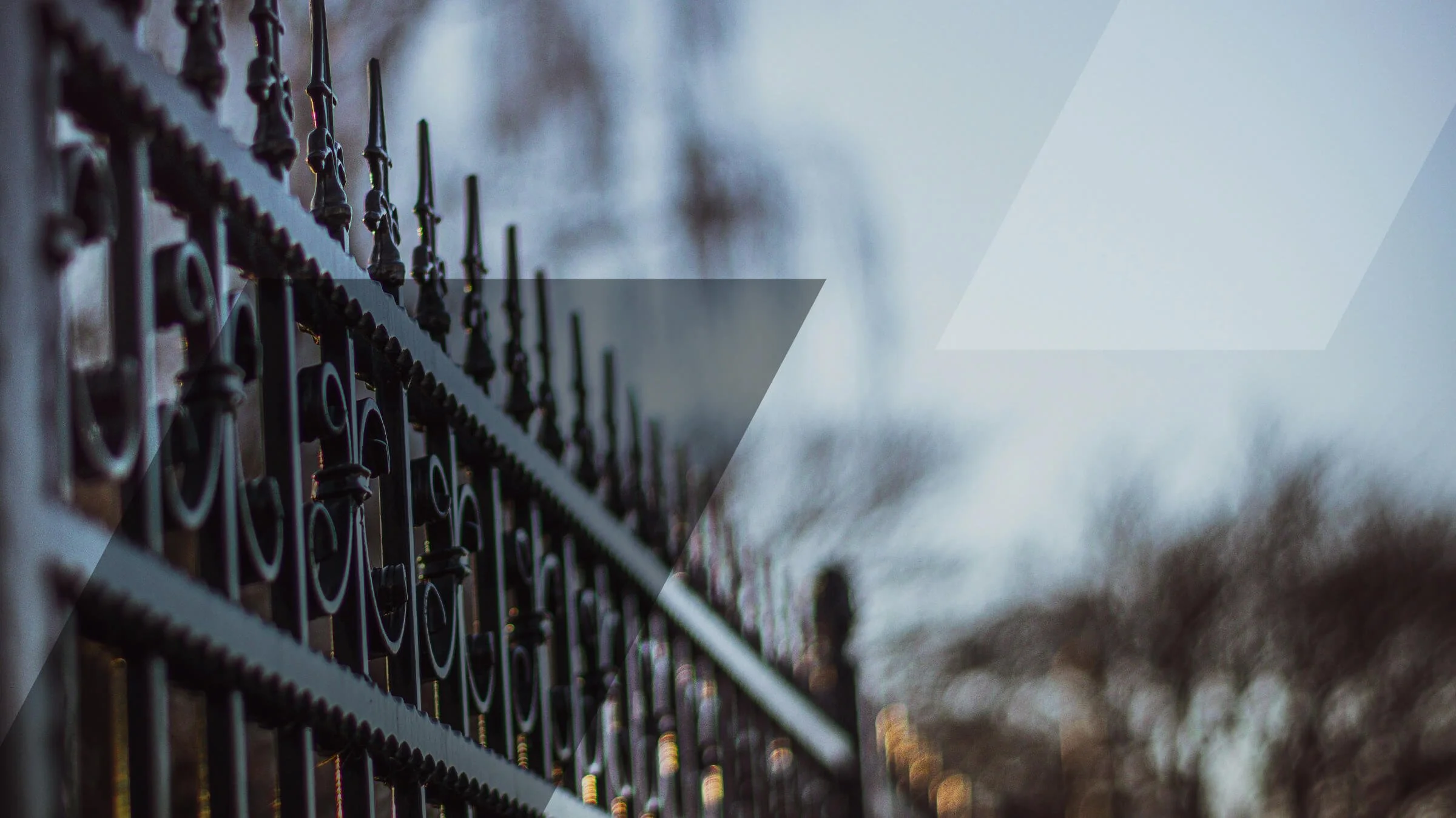 Ein schwarzer Metallzaun mit dekorativen Spitzen vor einem verschwommenen Hintergrund von Bäumen und Himmel, aufgenommen bei Dämmerung.