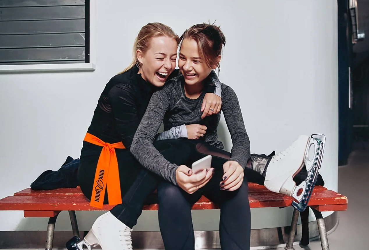 Figure skaters sitting at the rink bench wearing a neon Rink Ribbon™ before stepping onto the ice.