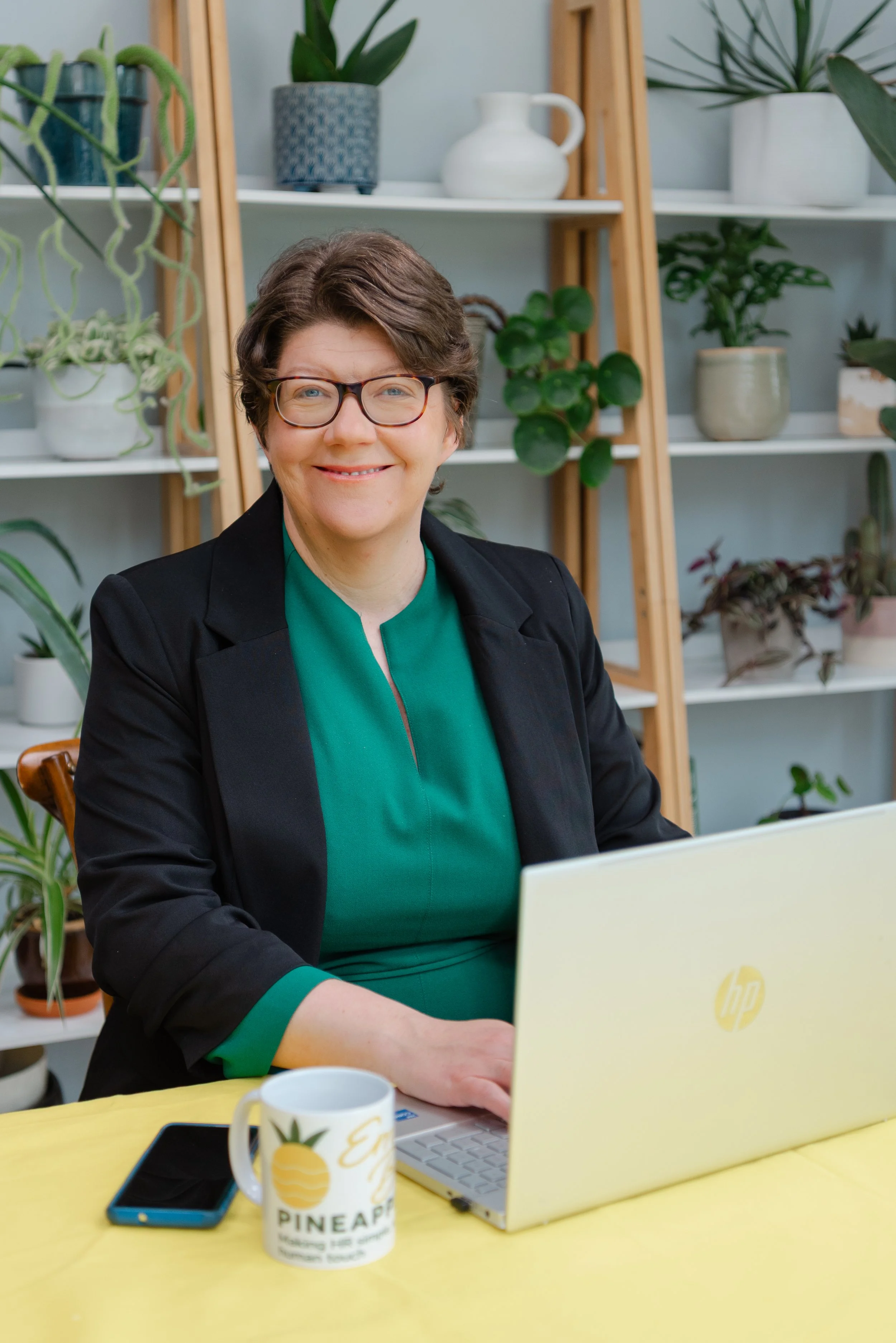 A woman with short brown hair, glasses, and a black blazer sitting at a yellow table with a laptop, a mug, and a phone in front of her, working in a room with shelves filled with green potted plants in the background.