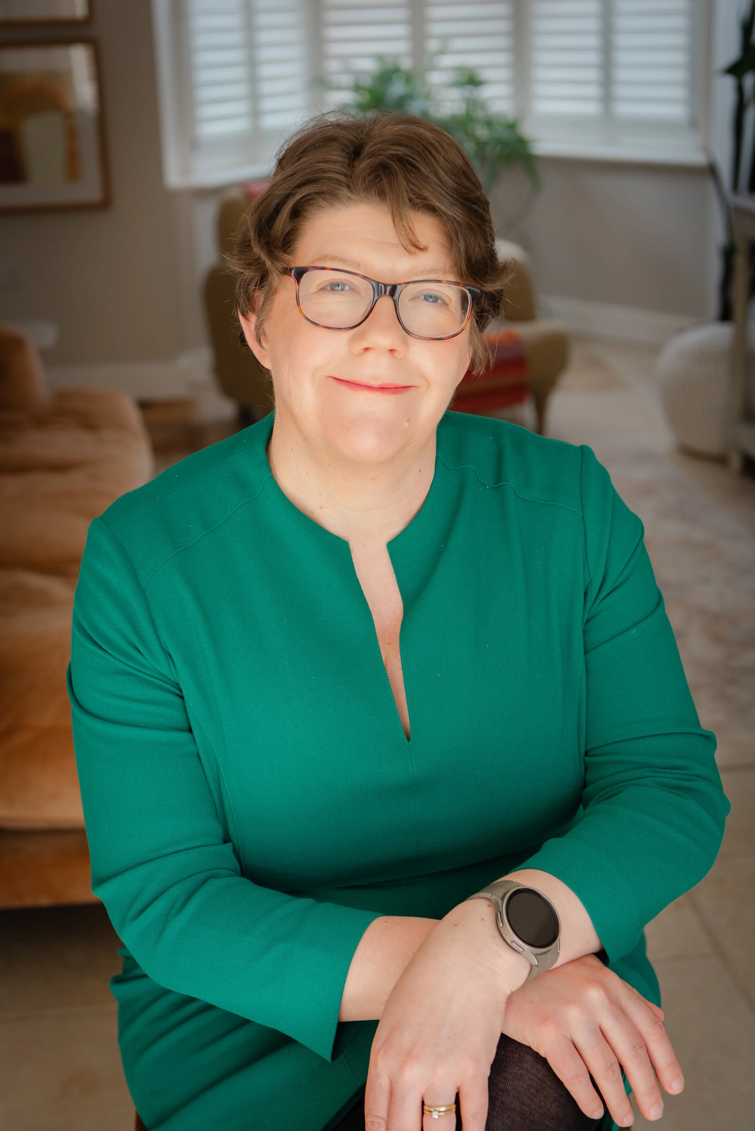 A woman with short brown hair, glasses, and a green top sitting indoors with a smile, with a plant and window blinds in the background.