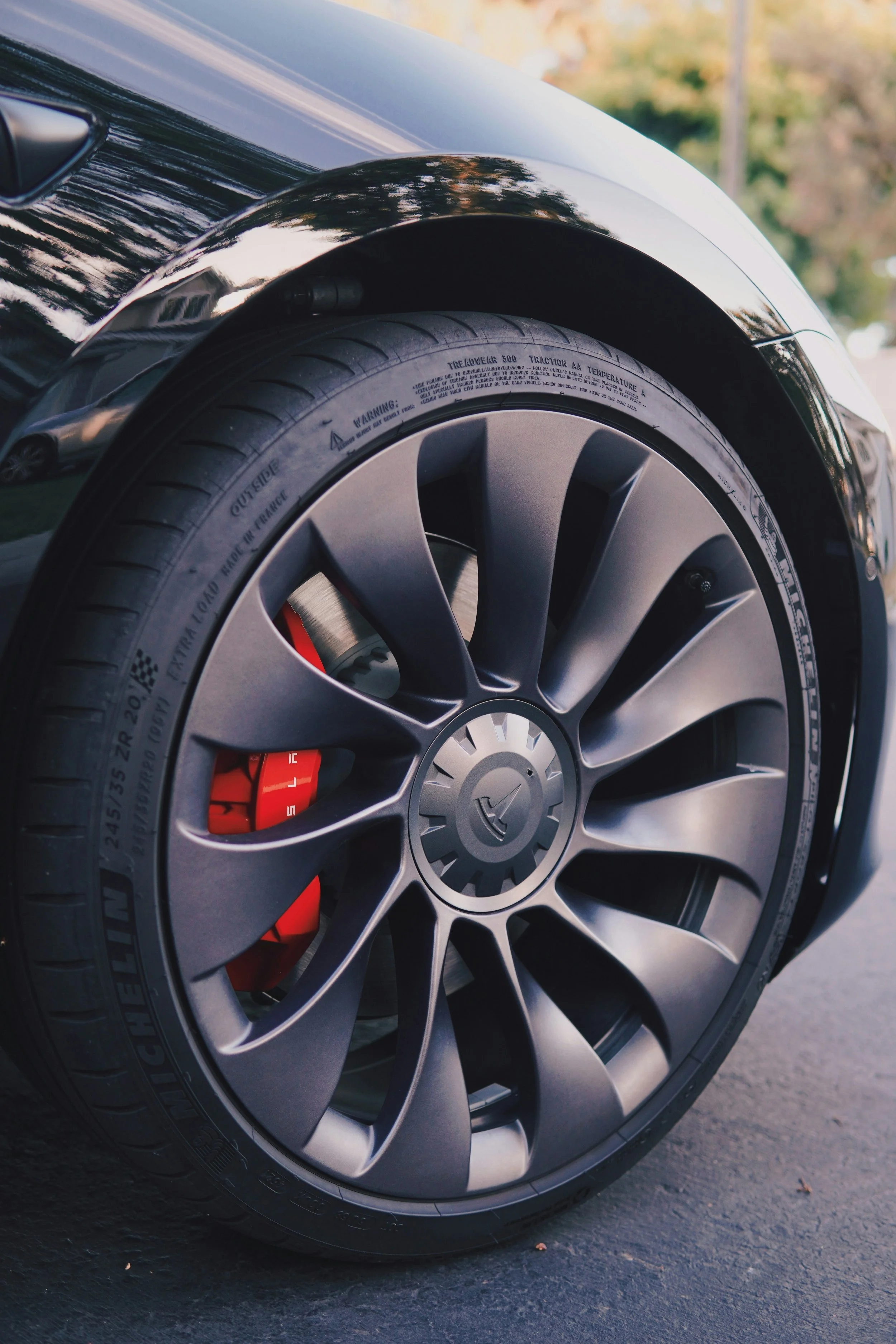 Close-up of a modern car wheel with a matte gray alloy rim, red brake caliper, and Michelin tire, parked on a paved surface with a blurred background.
