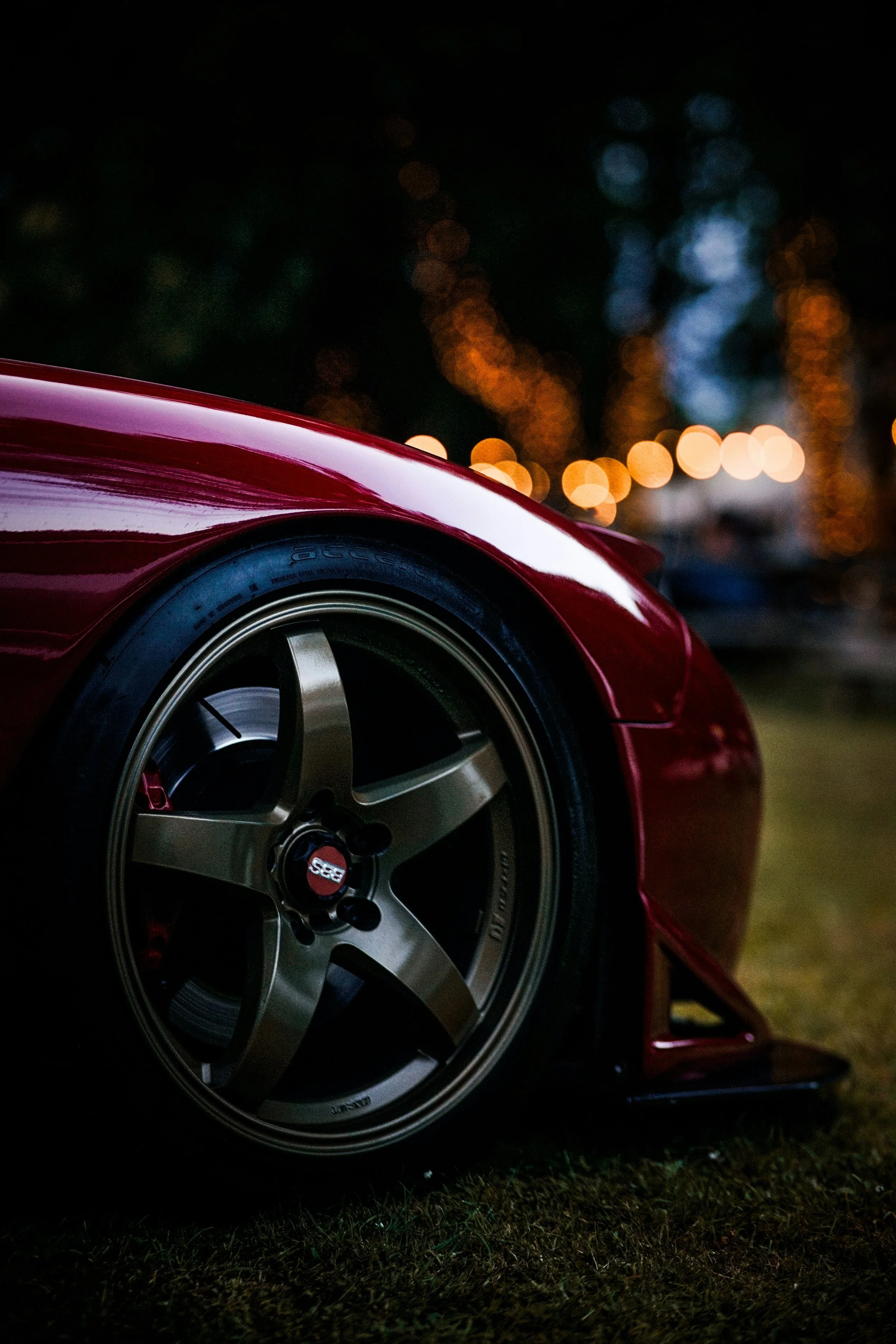 Close-up of a red sports car tire and wheel at night, with blurred, colorful bokeh lights in the background.