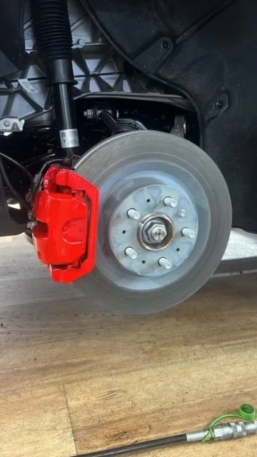 Close-up of a car's front wheel with a red brake caliper and a silver brake disc, showing suspension and brake components.