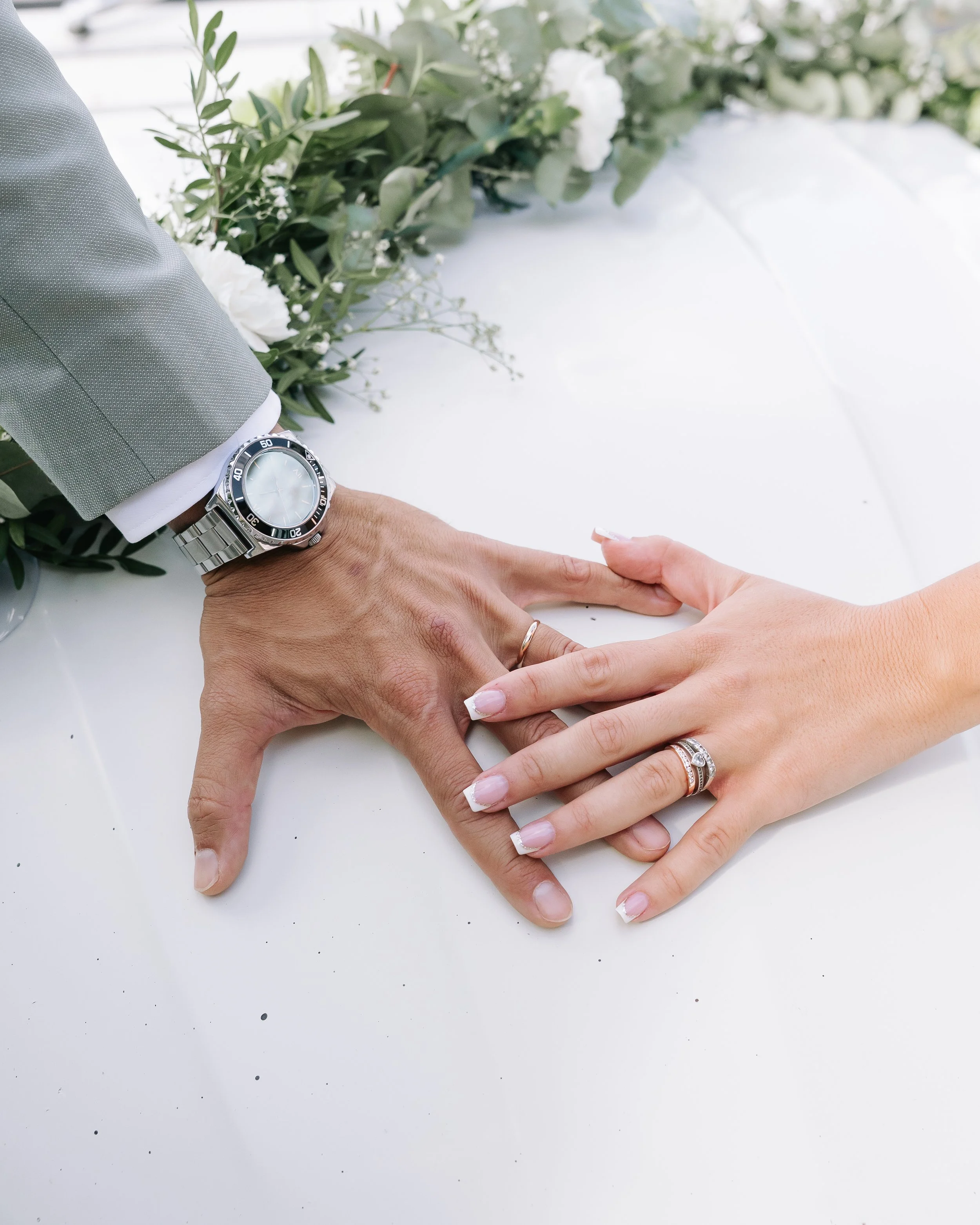 Deux mains, une homme portant une montre en acier et une femme portant plusieurs anneaux, tiennent la main de l'autre sur une table blanche. À l'arrière-plan, il y a une décoration florale avec des feuilles vertes et des fleurs blanches.