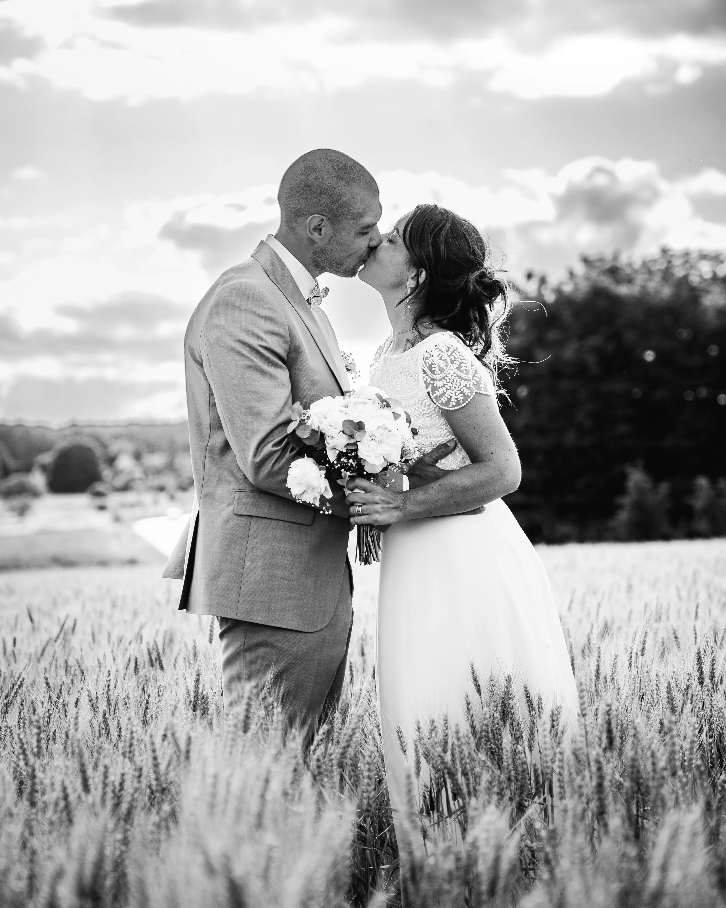 Un couple en tenue de mariage s'embrasse dans un champ de blé, en noir et blanc, avec un ciel nuageux en arrière-plan.