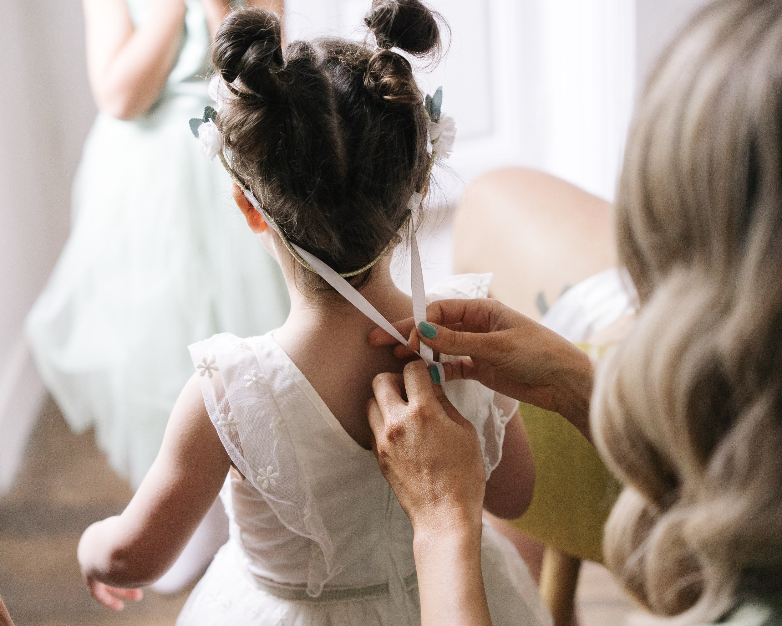 Une personne aide une jeune fille à enfiler un masque. La jeune fille porte une robe blanche avec des détails en fleurs et ses cheveux sont coiffés en deux buns.