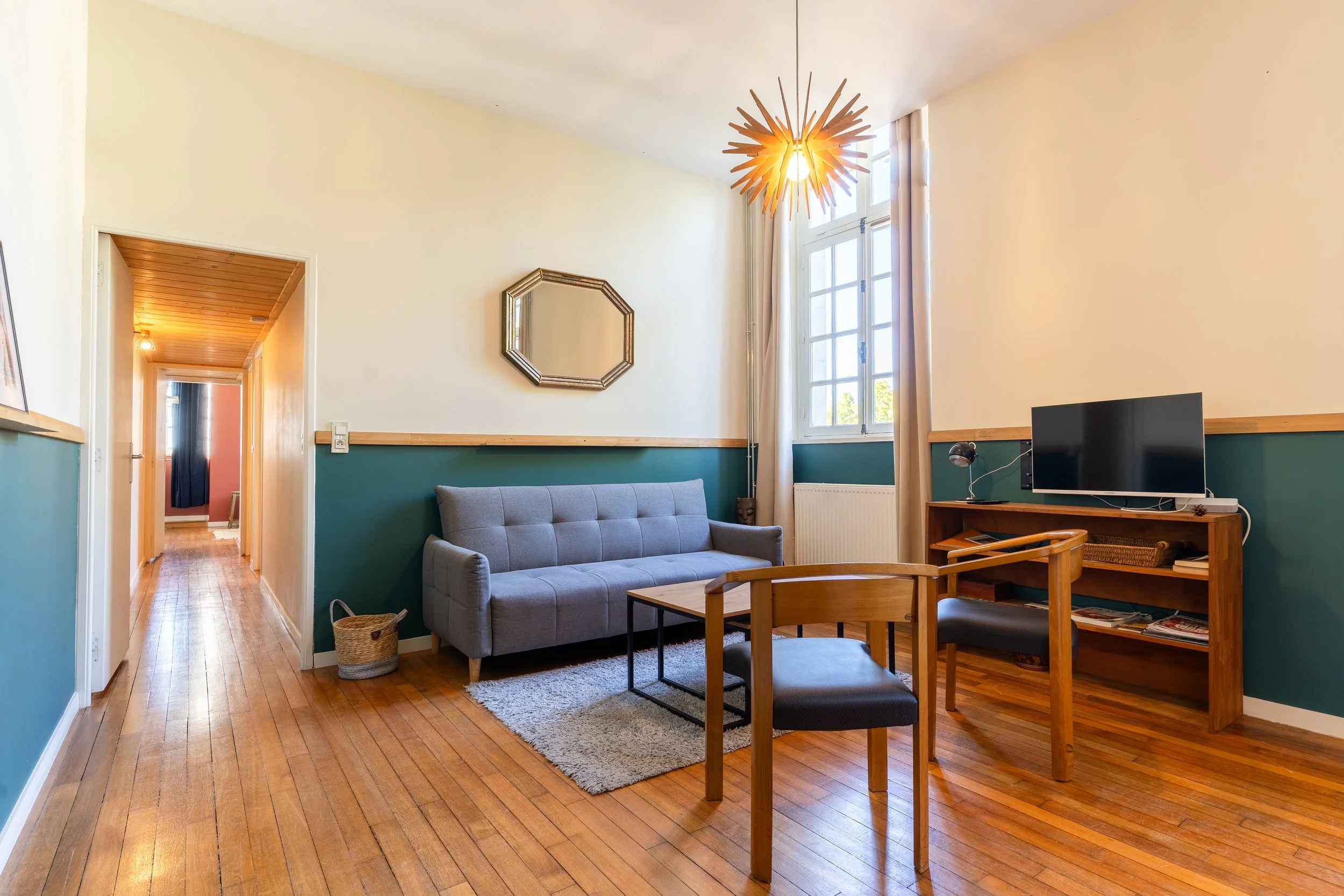 Salon avec canapé gris, table basse, télévision, étagère en bois, mur blanc avec un miroir, fenêtre avec rideaux, lampe suspendue en bois, sol en parquet, porte menant à un couloir avec plafond en bois.