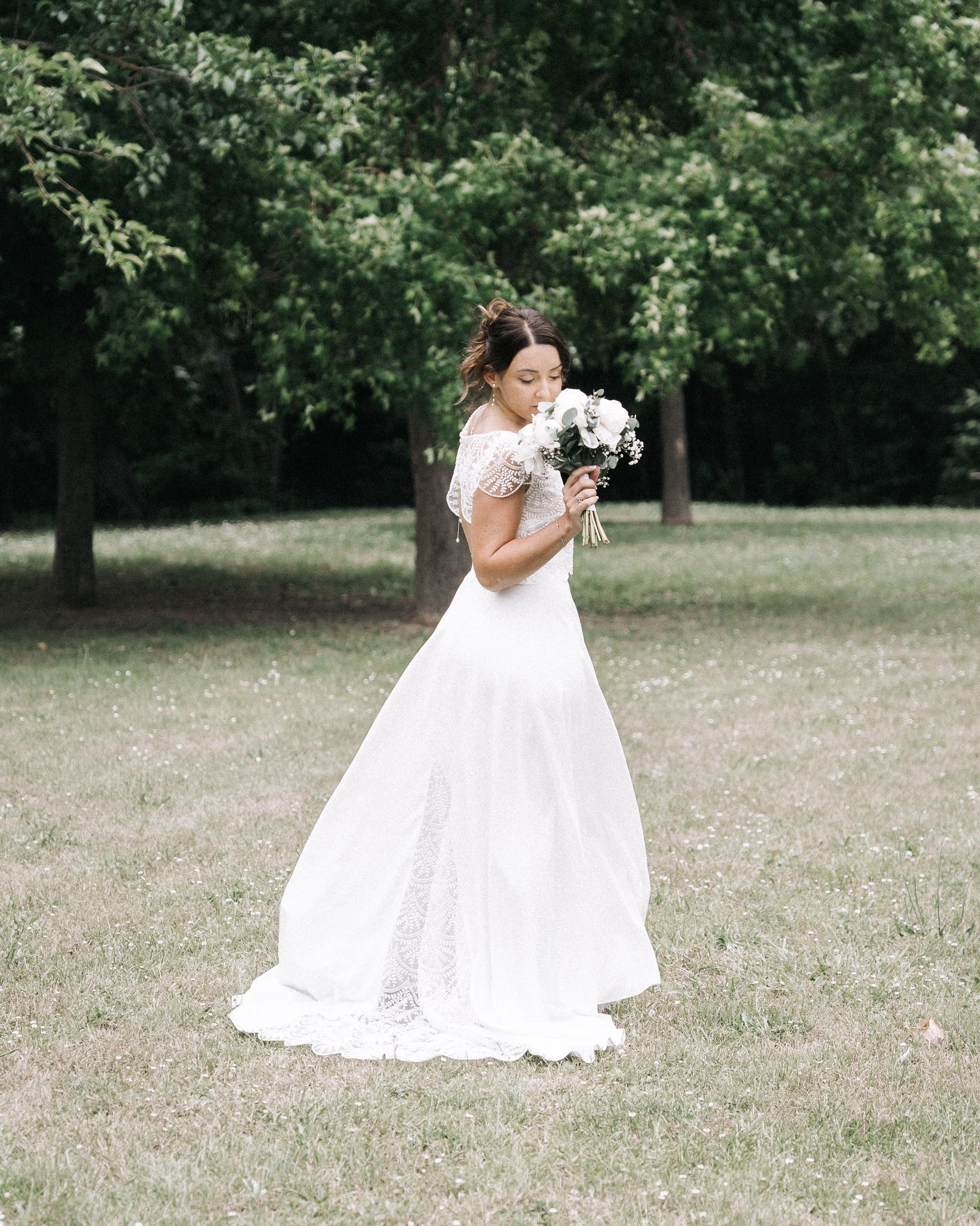 Jeune femme en robe de mariée blanche tenant un bouquet de fleurs, debout dans un parc avec des arbres en arrière-plan.