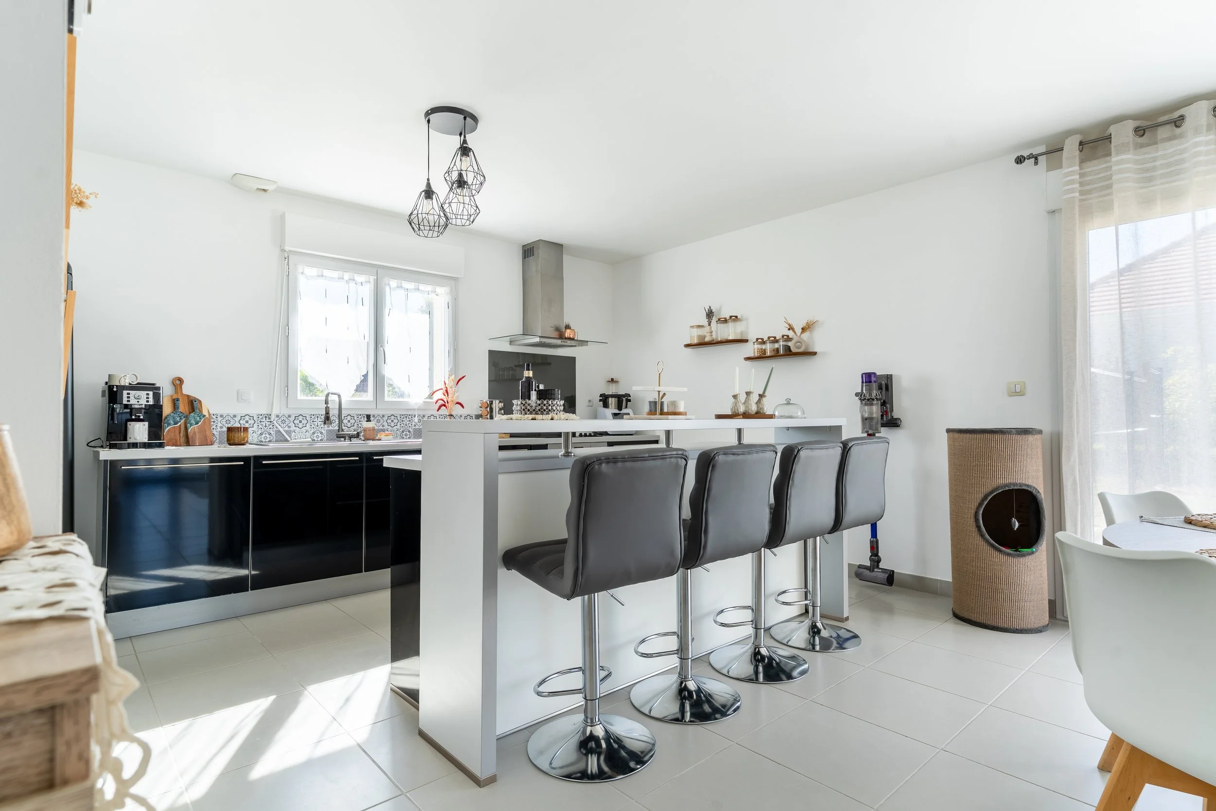 Cuisine moderne avec un ilot central, chaises hautes en cuir noir, lampes suspendues en métal, grande fenêtre avec rideaux, et murs blancs décorés avec des étagères en bois.