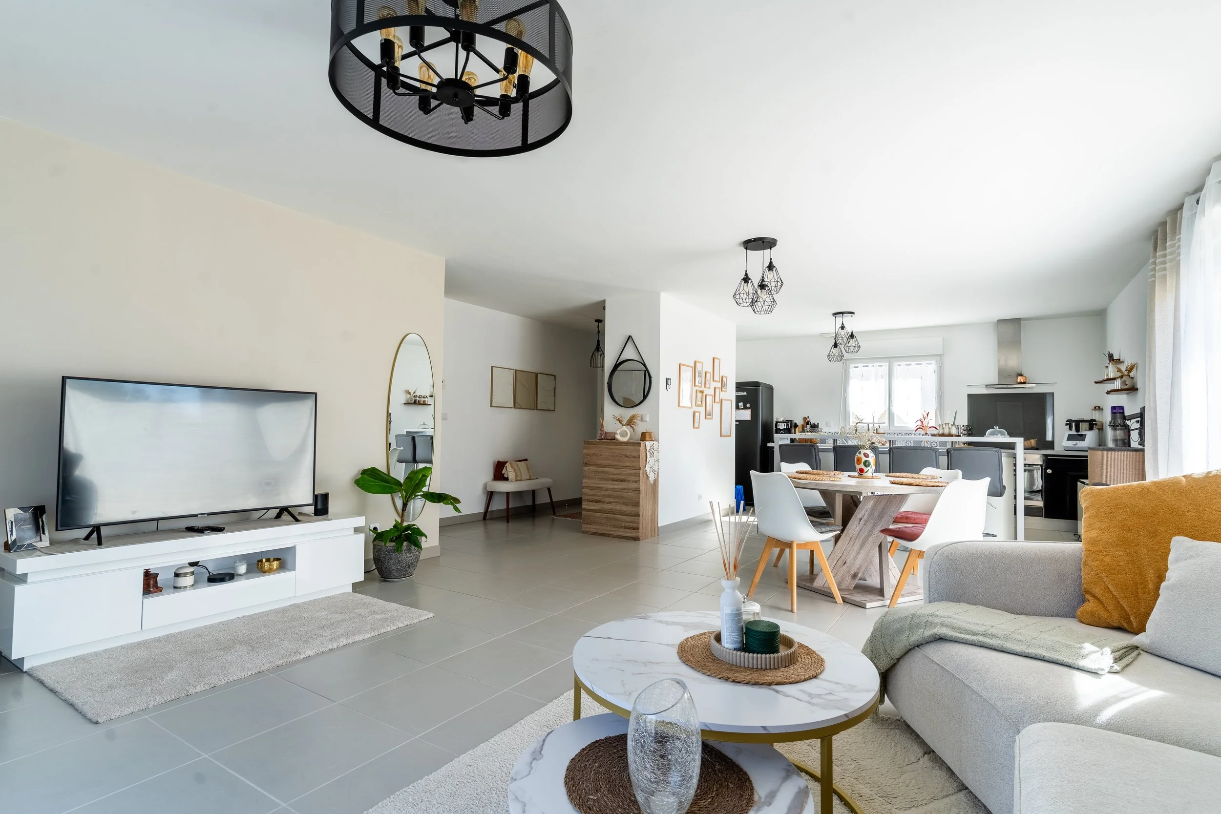 Salon moderne et lumineux avec canapé gris, table basse en marbre, télévision à écran plat, plantes vertes, et décoration minimaliste. Salle à manger attenante avec table ronde et chaises blanches, cuisine en arrière-plan avec électroménagers et lumi