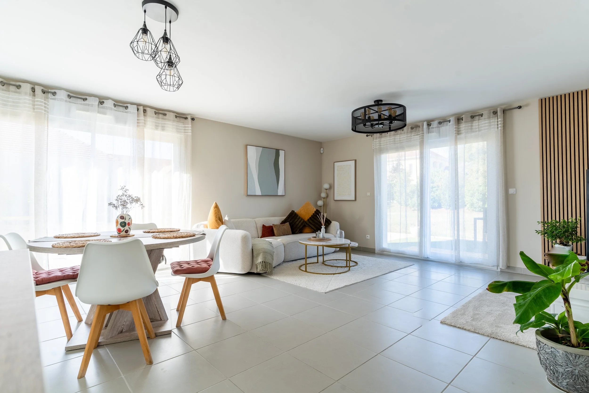 Salon moderne avec canapé beige, poufs, table basse ronde, tableaux, et grande fenêtre avec rideaux transparents.