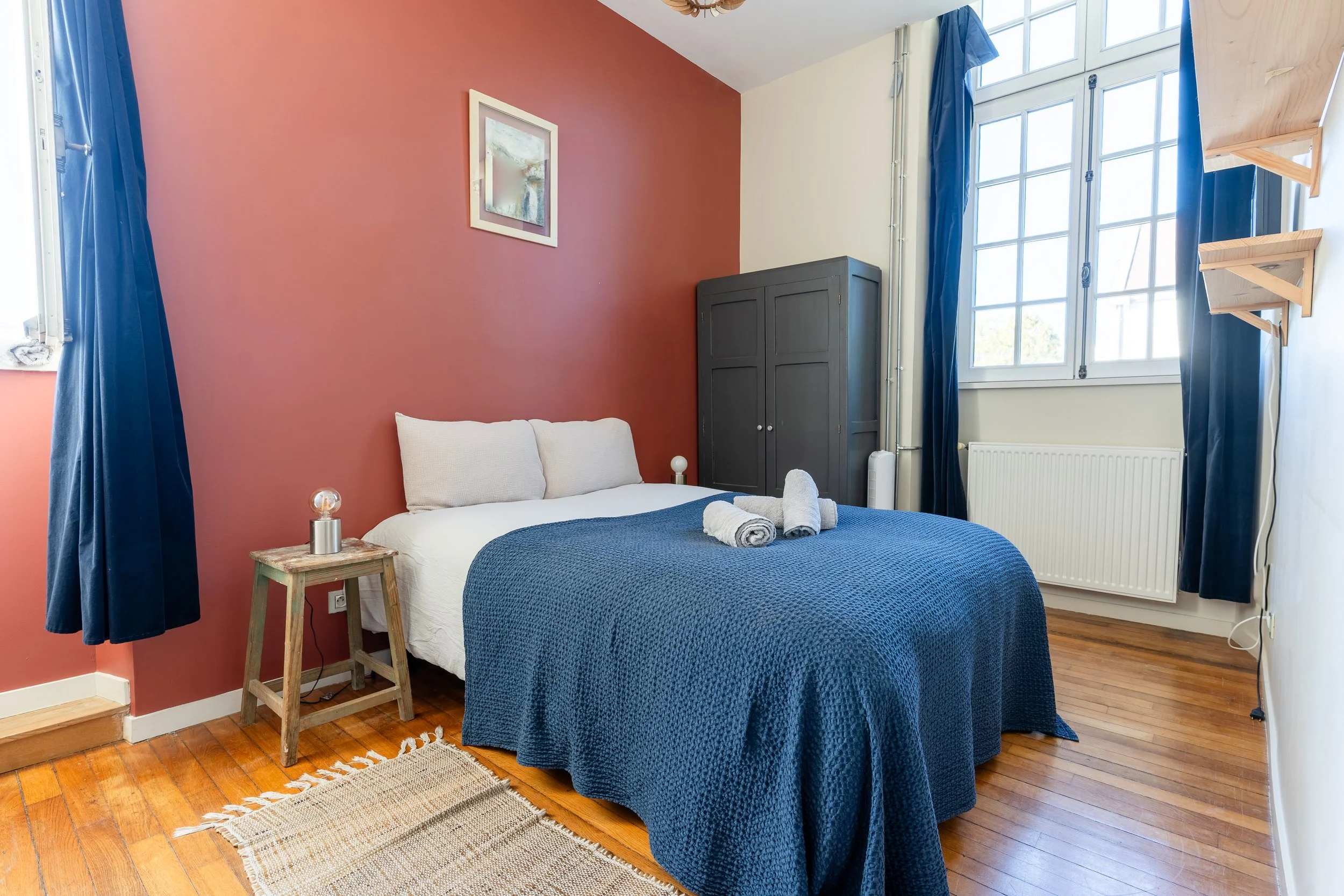 Chambre avec un lit fait avec une couverture bleue, deux serviettes pliées, une table de chevet en bois, une armoire noire, un tableau décoratif, un radiateur, des fenêtres avec rideaux bleus, et un sol en bois.