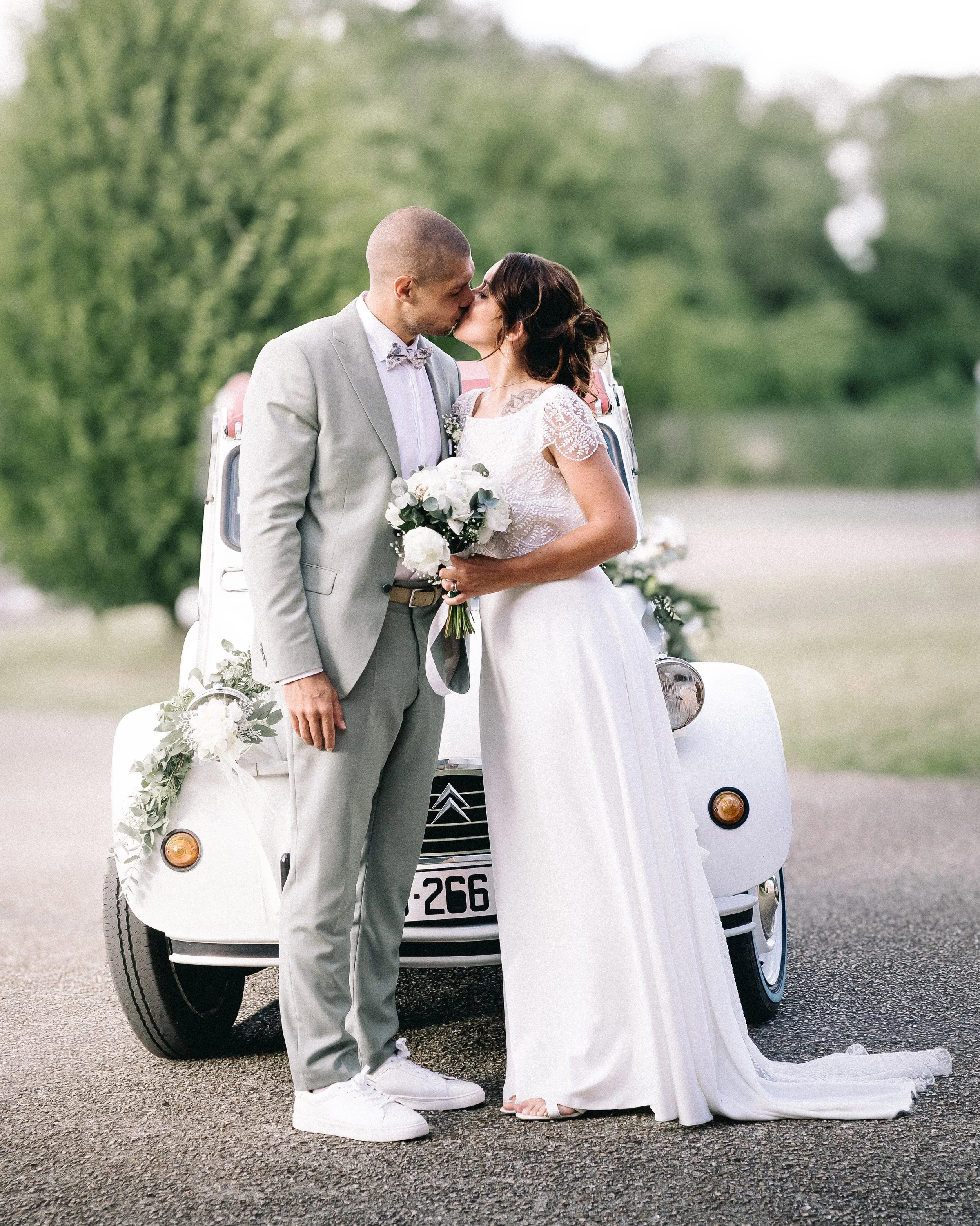 Un couple marié, un homme en costume gris et une femme en robe de mariage blanche, s'embrassent devant une petite voiture blanche décorée de fleurs, dans un environnement extérieur verdoyant.
